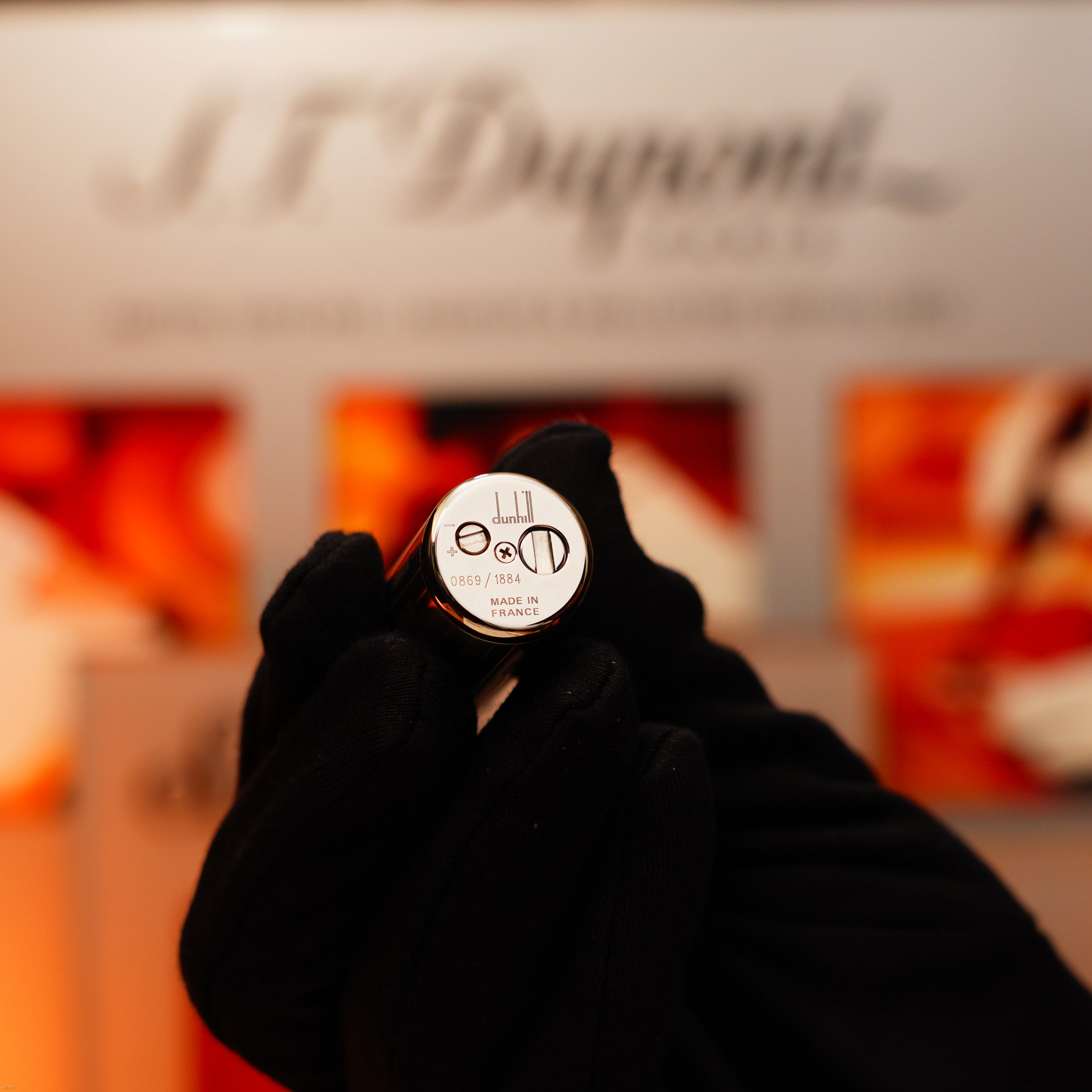 A black-gloved hand holds a vintage Alfred Dunhill Full Set GMT solid stainless steel lighter by Lucy Lighter, engraved with symbols and numbers; blurred branding appears in the background.