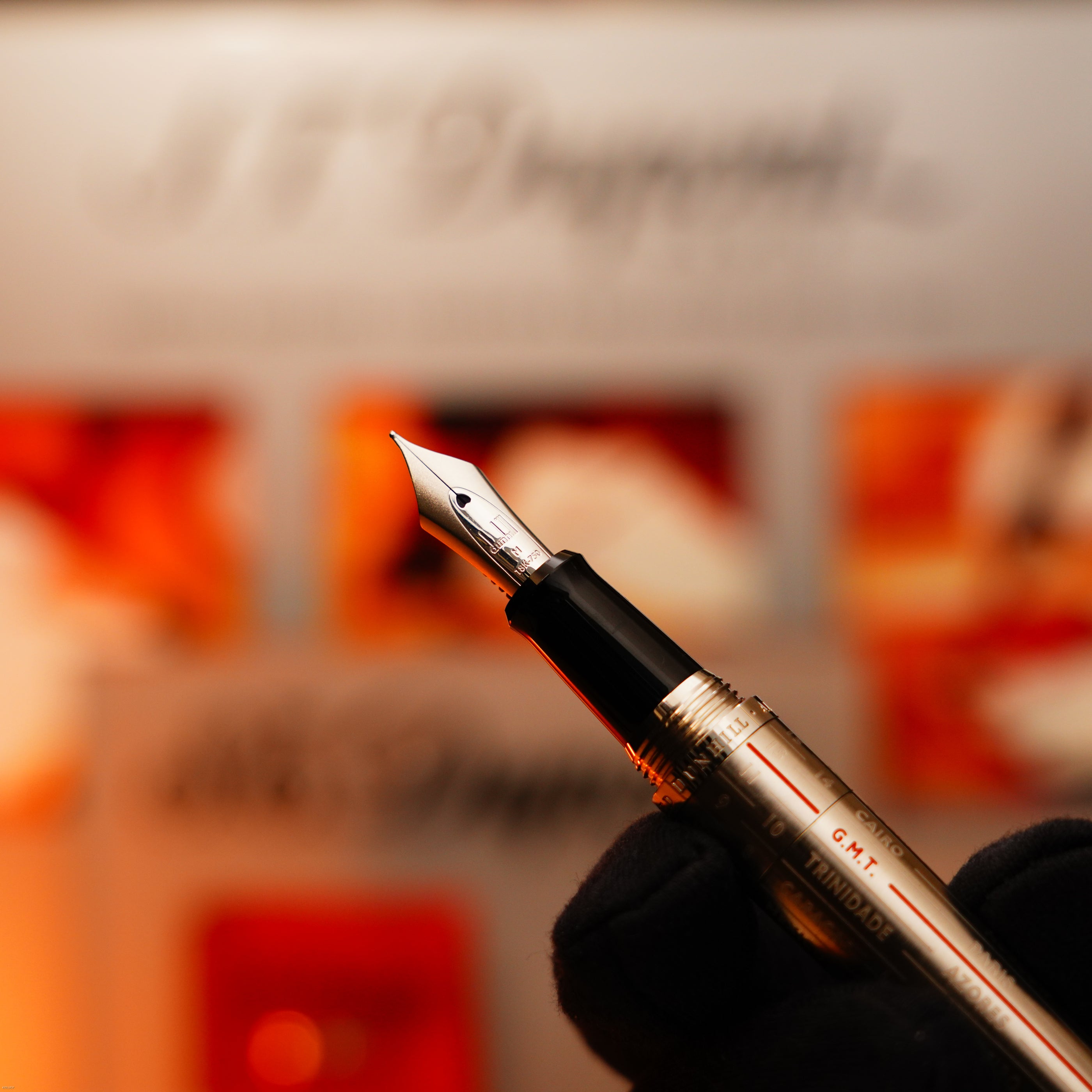 A gloved hand holds a Vintage Extremely Rare Alfred Dunhill Full Set GMT Full Solid Stainless Steel lighter by Lucy Lighter, shown in sharp focus against a blurred orange and white background—reflecting its assured quality.