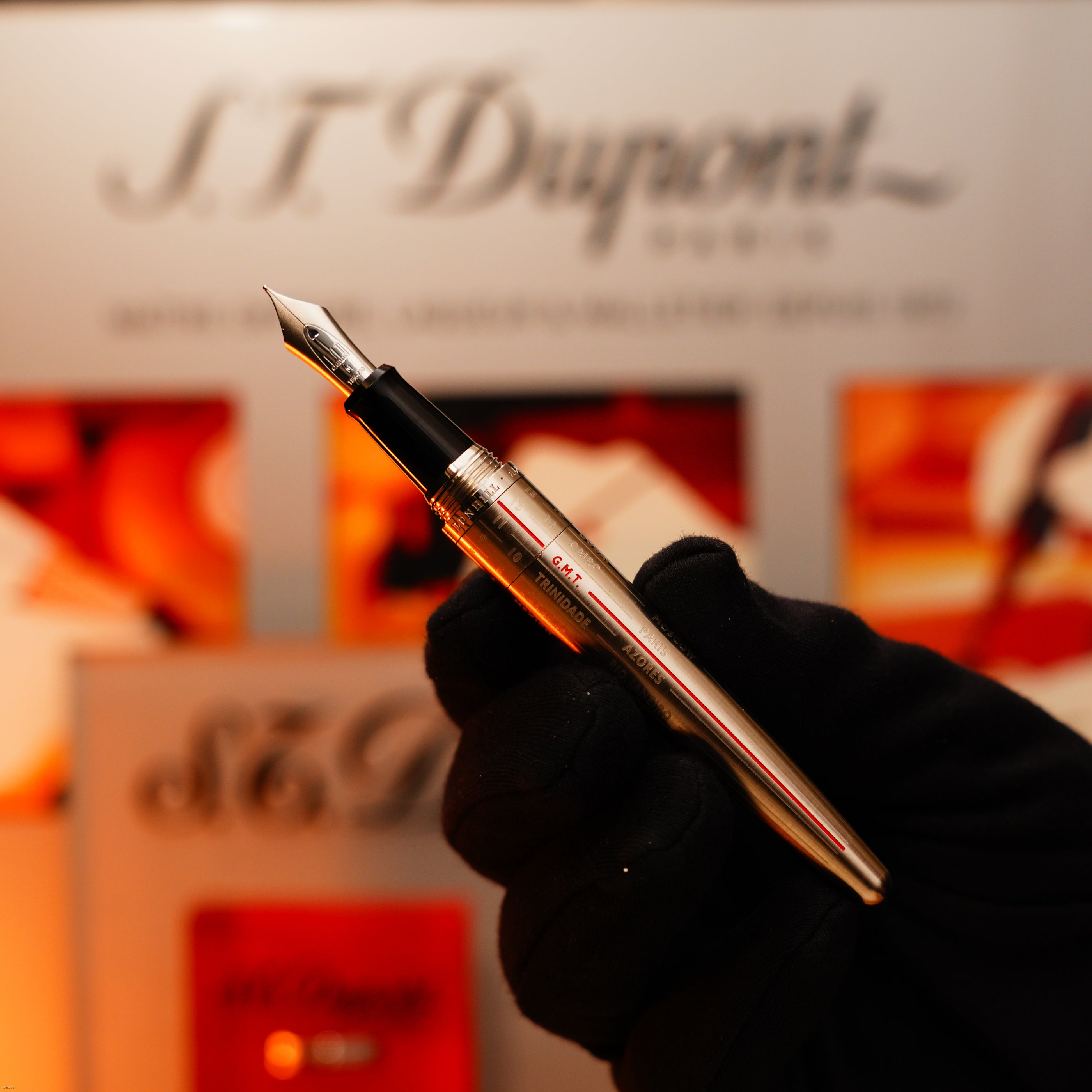 A gloved hand holds a rare, full set Alfred Dunhill GMT stainless steel lighter by Lucy Lighter, with a blurred background featuring the Alfred Dunhill signature set and packaging—highlighting the brand’s reputation for vintage luxury.