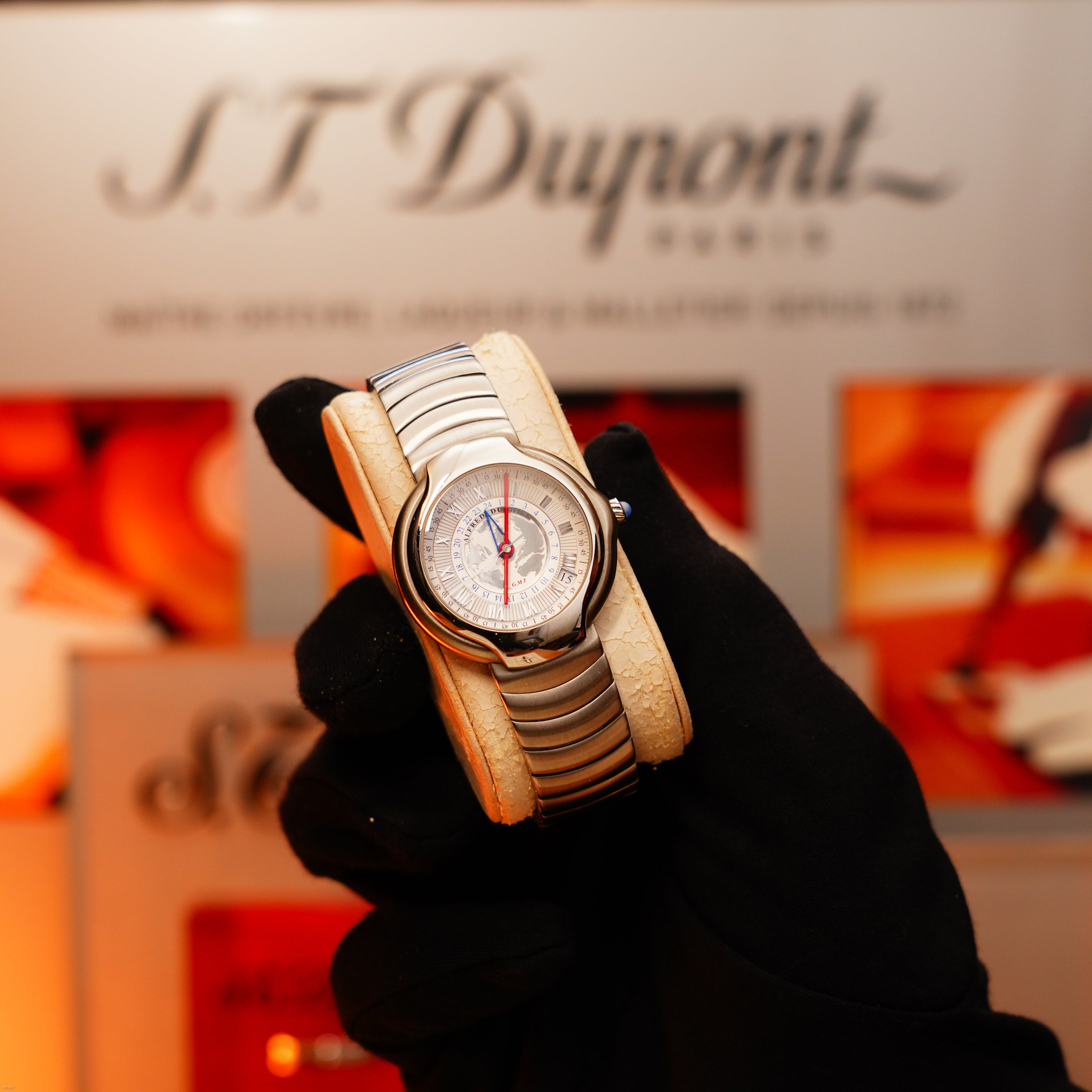 A gloved hand holds a Vintage Extremely Rare Alfred Dunhill GMT Full Solid Stainless Steel watch from Lucy Lighter, showcased before a blurred display—emphasizing the full set's quality and exclusivity.