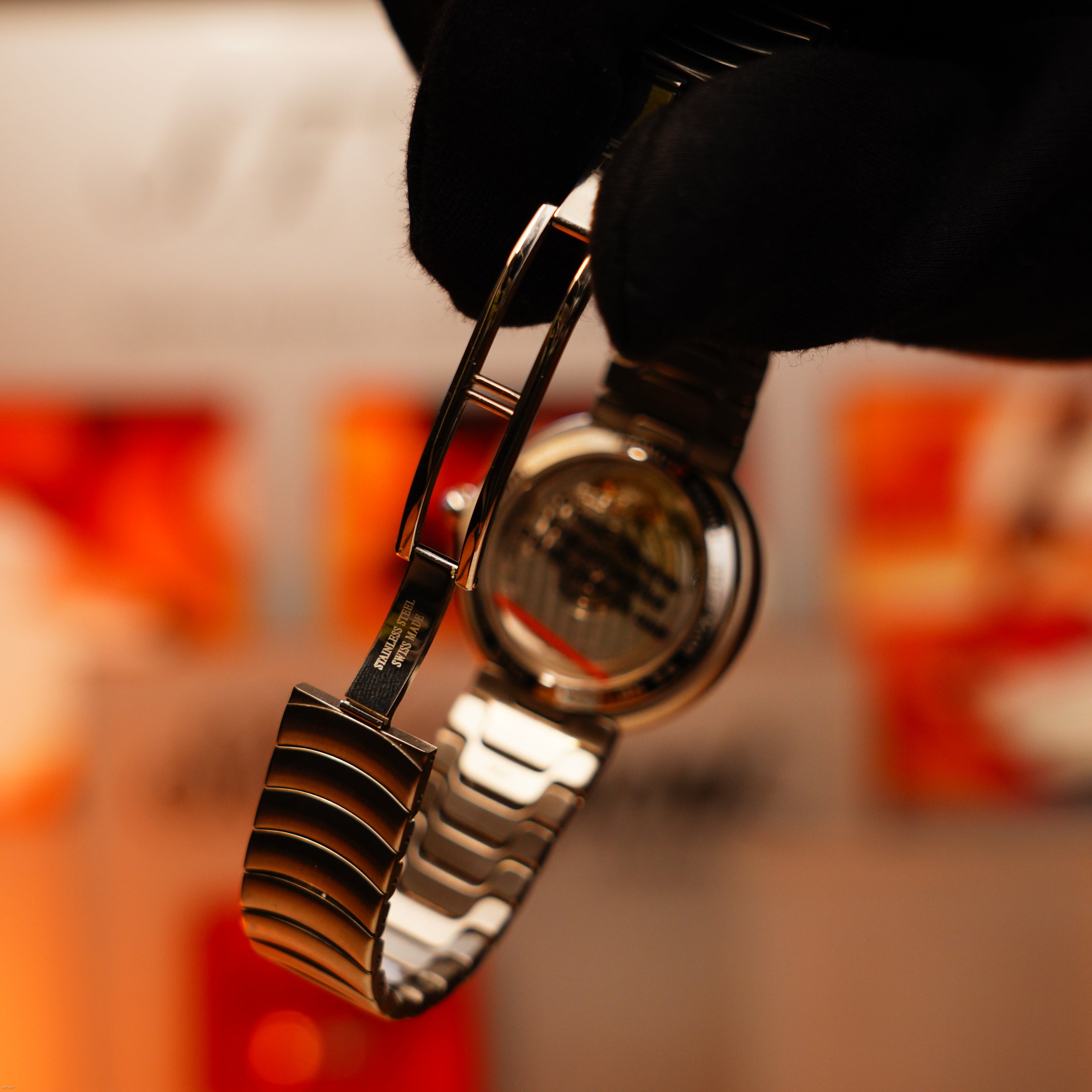 A gloved hand holds the Vintage Extremely Rare Alfred Dunhill GMT Full Solid Stainless Steel watch by Lucy Lighter, showing the back and clasp against a blurred orange-white background—a must-have for collectors seeking authentic full sets.