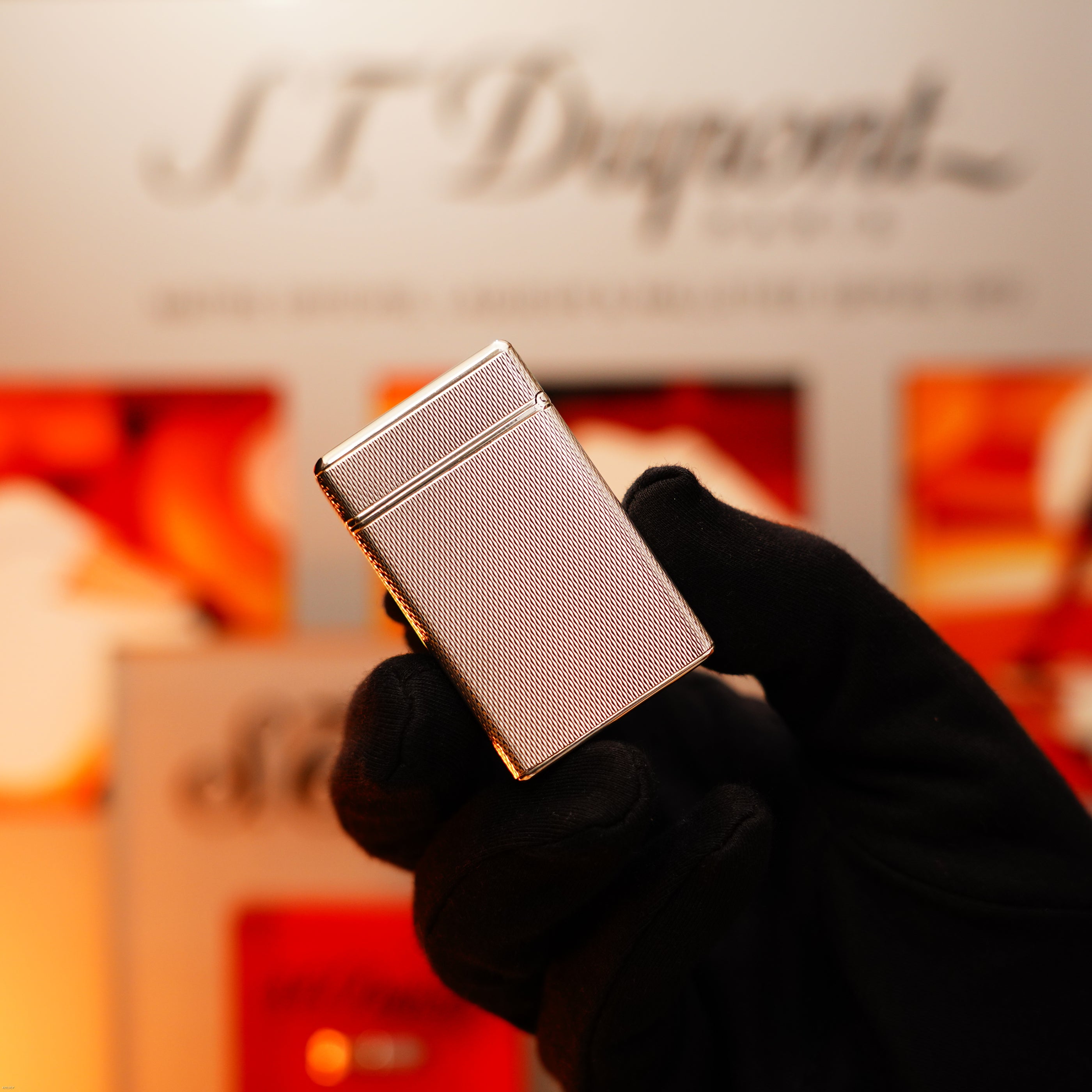 A gloved hand holds the Vintage 1970 Extremely Rare 1/1 De St Dupont Solid 925 Sterling Ligne 1 Heavy Barley Pattern Lighter by Lucy Lighter, with a blurred background highlighting the iconic logo and product displays.