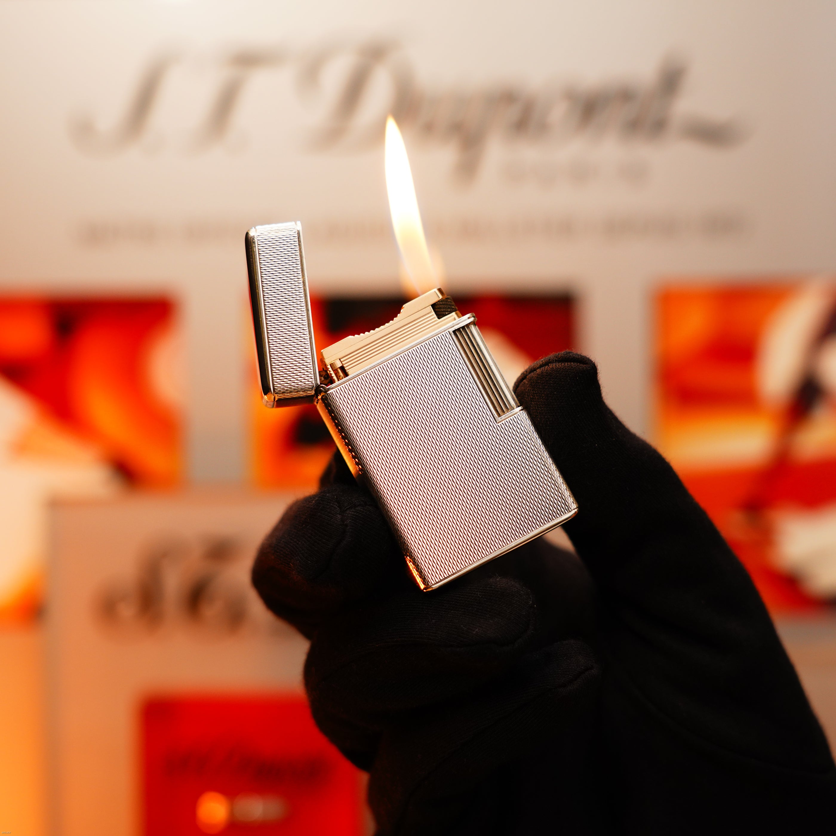 A gloved hand holds the Vintage 1970 Extremely Rare 1/1 De St Dupont Solid 925 Sterling Rare Ligne 1 Heavy Barley Pattern Lighter by Lucy Lighter, its lit flame and blurred branded background highlighting luxury and rarity.