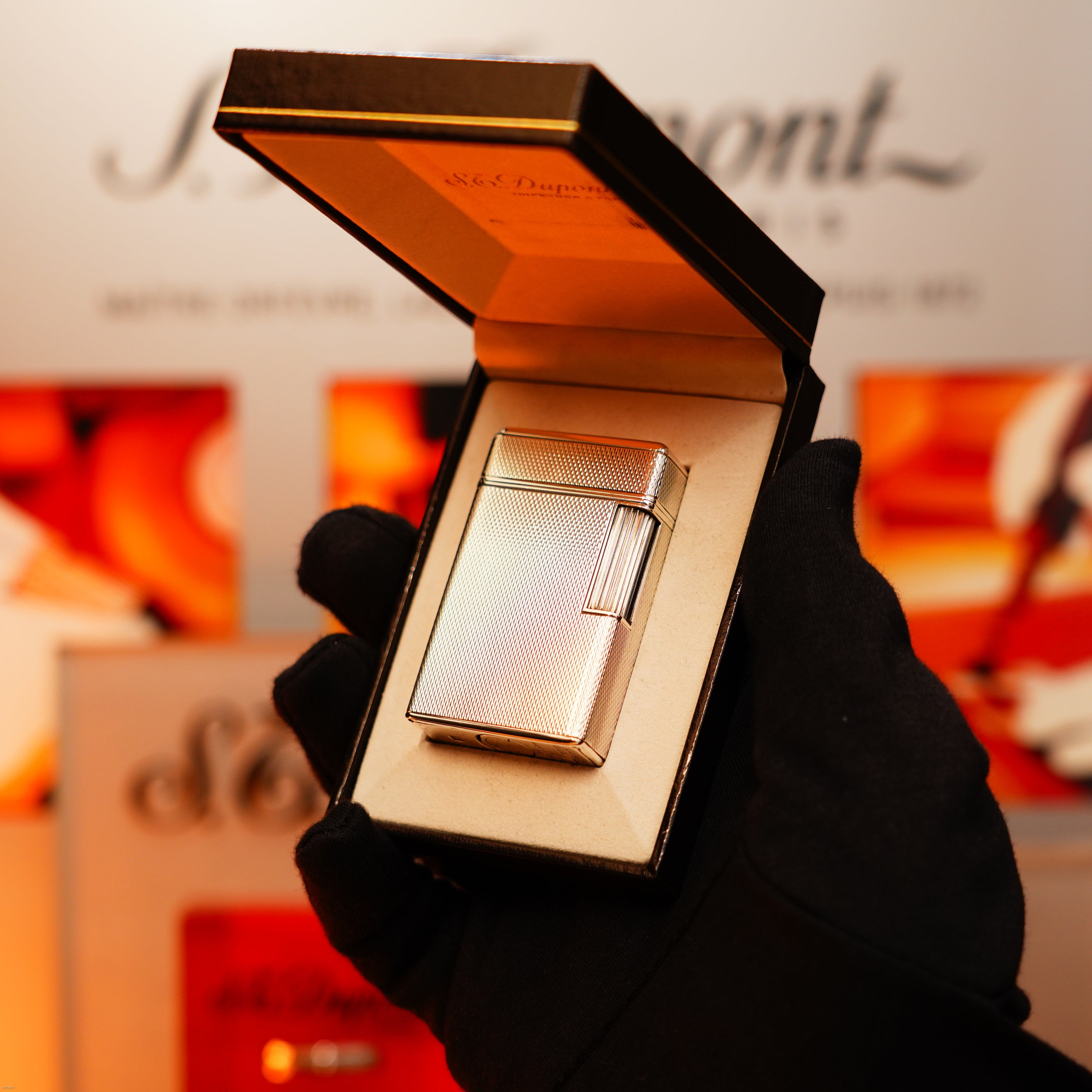 A gloved hand holds an open box revealing the Vintage 1970 Extremely Rare 1/1 De St Dupont Solid 925 Sterling Ligne 1 Heavy Barley Pattern Lighter by Lucy Lighter, set against a blurred background with brand signage, exuding timeless elegance.