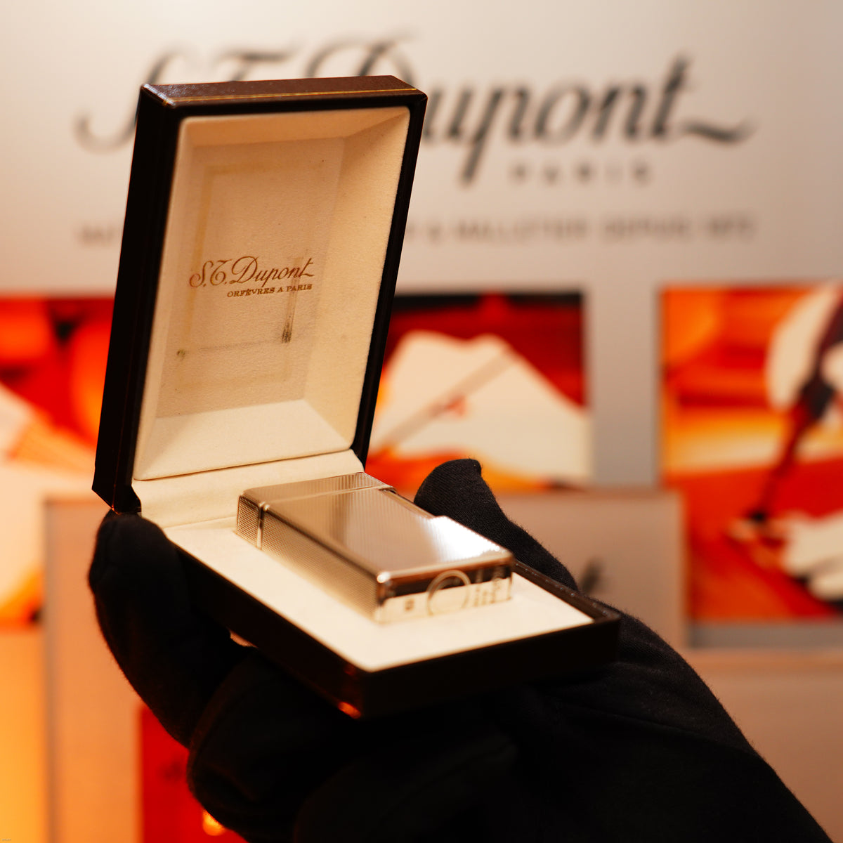 A gloved hand holds an open Lucy Lighter box showcasing the Vintage 1970 Extremely Rare 1/1 De St Dupont Solid 925 Sterling Ligne 1 Heavy Barley Pattern Lighter, with a blurred sign in the background.