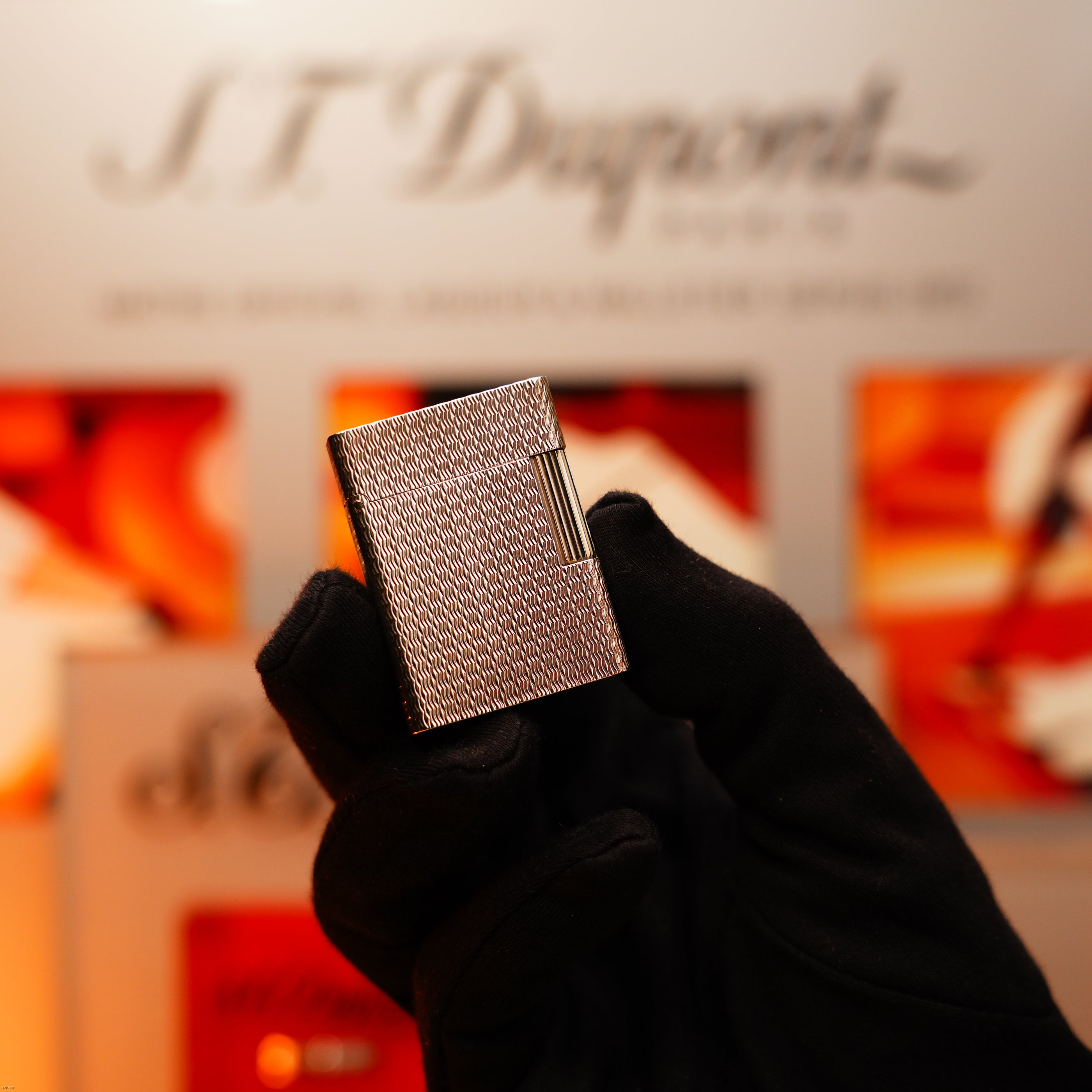A gloved hand holds the Vintage 1970 Extremely Rare 1/1 De St Dupont Solid 925 Sterling Rare Ligne 1 Heavy Guilloché Pattern Lighter by Lucy Lighter, shown against a blurred background with the S.T. Dupont logo and colorful displays.