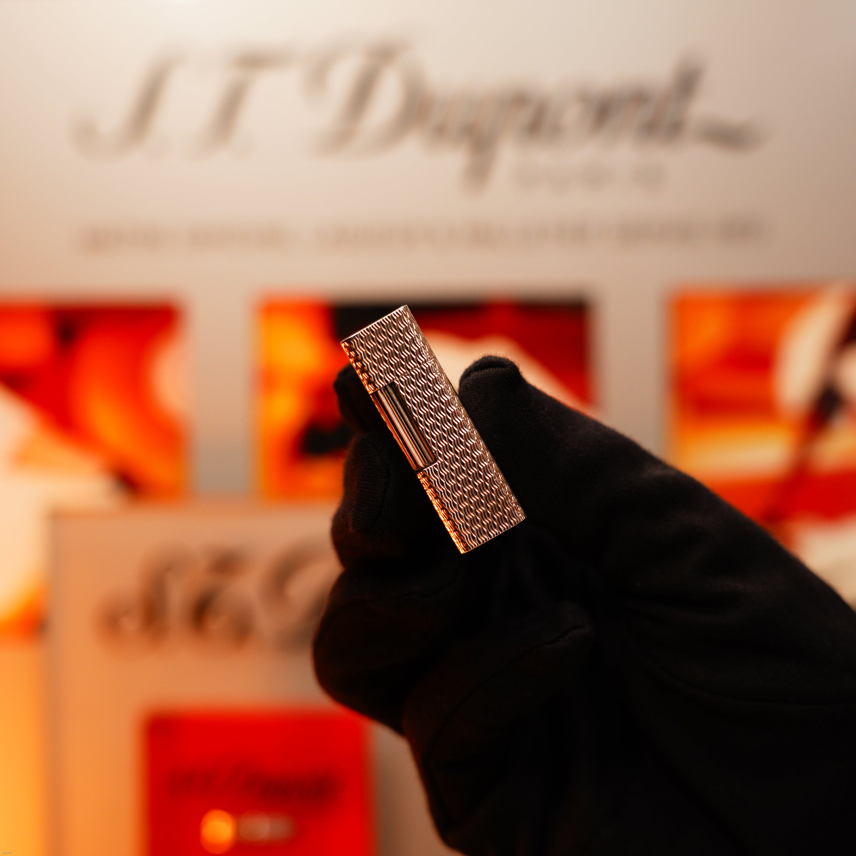 A gloved hand holds the Vintage 1970 Extremely Rare 1/1 De St Dupont Solid 925 Sterling Rare Ligne 1 Heavy Guilloché Pattern Lighter by Lucy Lighter, with blurred branding and packaging in the background, evoking luxury and exclusivity.