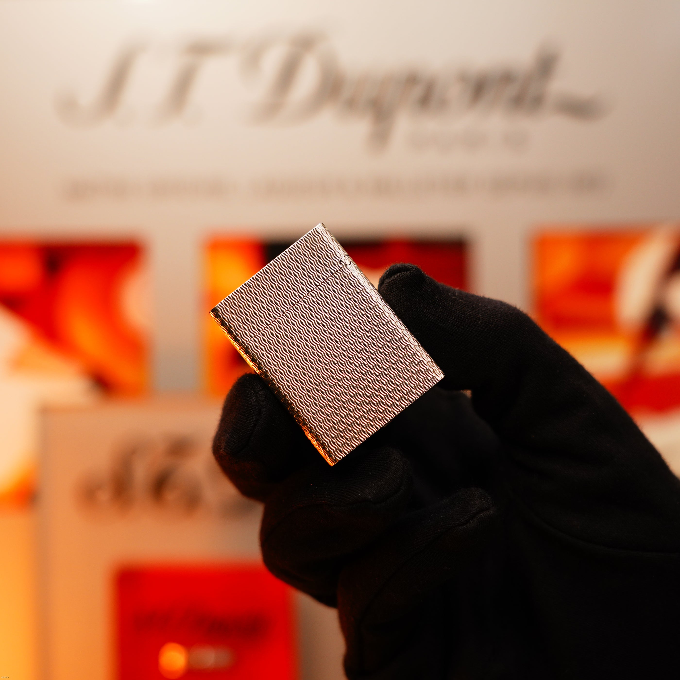 A gloved hand holds the Vintage 1970 Extremely Rare 1/1 De St Dupont Solid 925 Sterling Rare Ligne 1 Heavy Guilloché Pattern Lighter by Lucy Lighter, its textured design radiating luxury with blurred branding in the background.