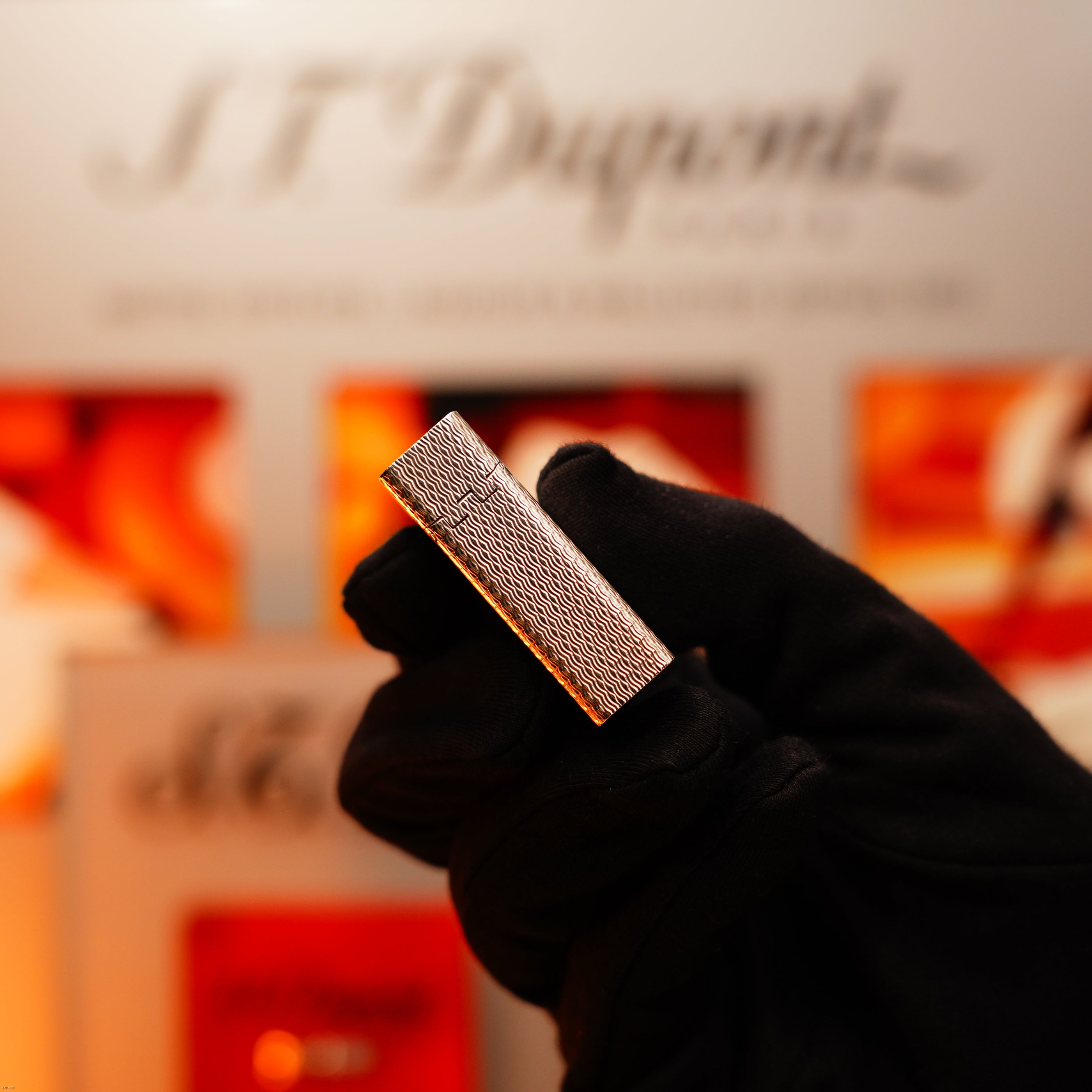 A gloved hand holds the Lucy Lighter Vintage 1970 Extremely Rare 1/1 De St Dupont Solid 925 Sterling Ligne 1 Heavy Guilloché Pattern Lighter before a blurred S.T. Dupont display.