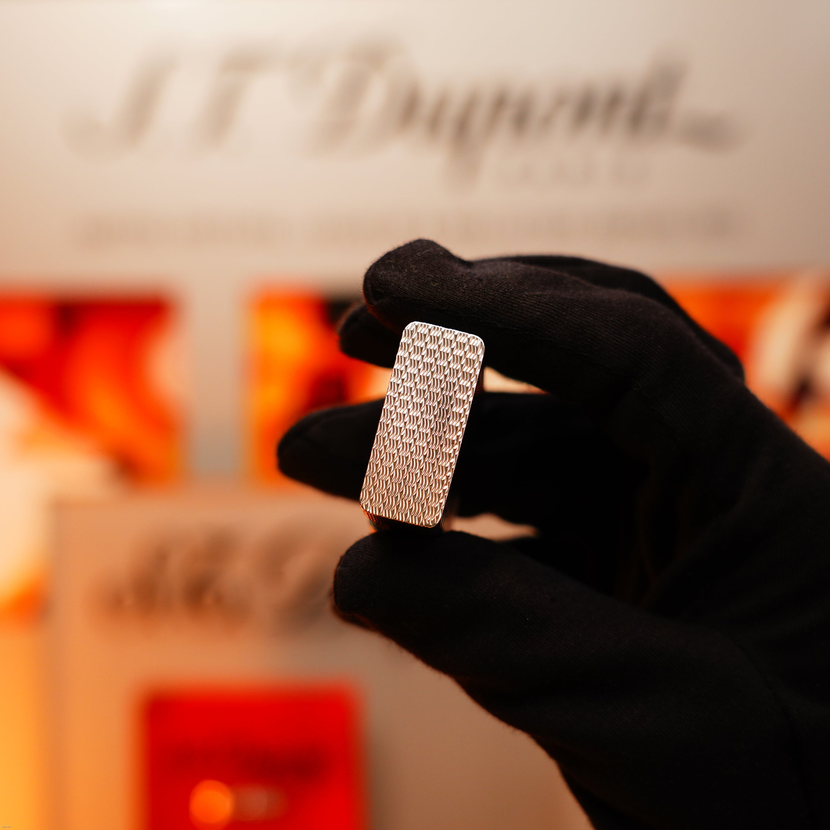A gloved hand holds a Lucy Lighter Vintage 1970 Extremely Rare 1/1 De St Dupont Solid 925 Sterling Ligne 1 Heavy Guilloché Pattern lighter, with a blurred S.T. Dupont sign in the background.