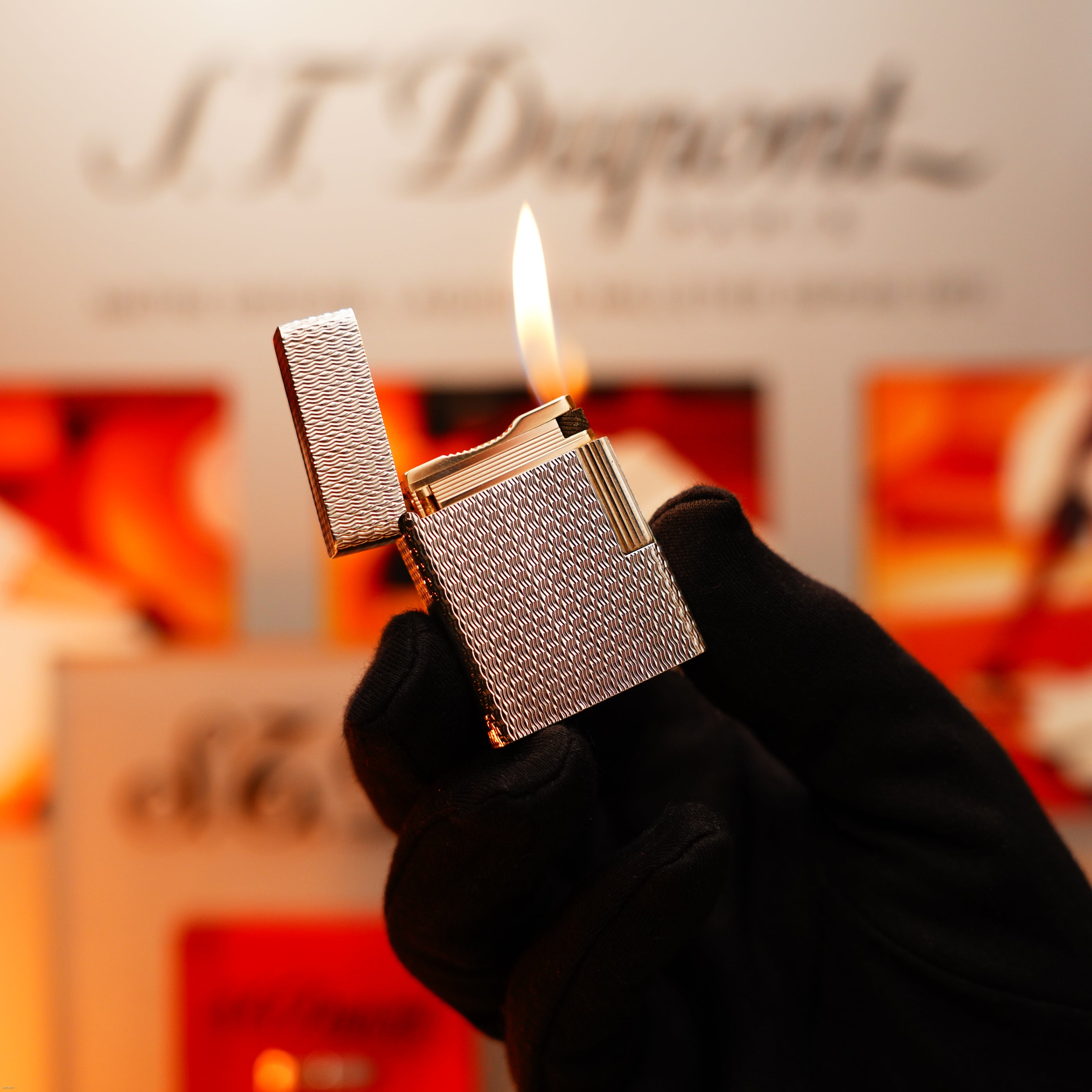 A hand in a black glove holds the Lucy Lighter Vintage 1970 Extremely Rare 1/1 De St Dupont Solid 925 Sterling Ligne 1 Heavy Guilloché Pattern Lighter, lit and open, with a blurred background and showcasing its 12-month warranty.