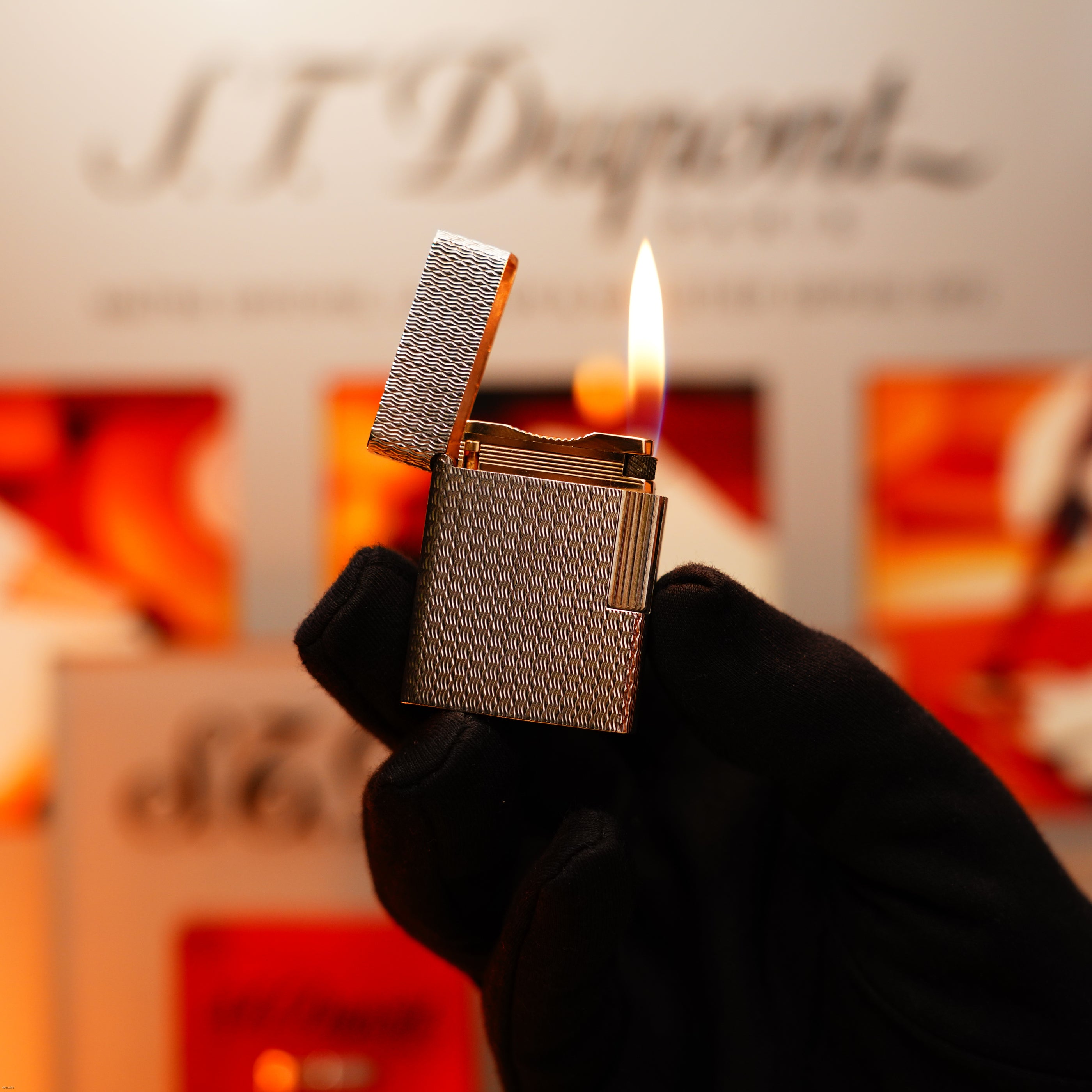 A gloved hand holds a lit Lucy Lighter Vintage 1970 Rare Ligne 1 925 Sterling Guilloché Pattern Lighter with open lid; blurred, warm-toned background highlights its luxury and exclusivity. Comes with a 12-month warranty.
