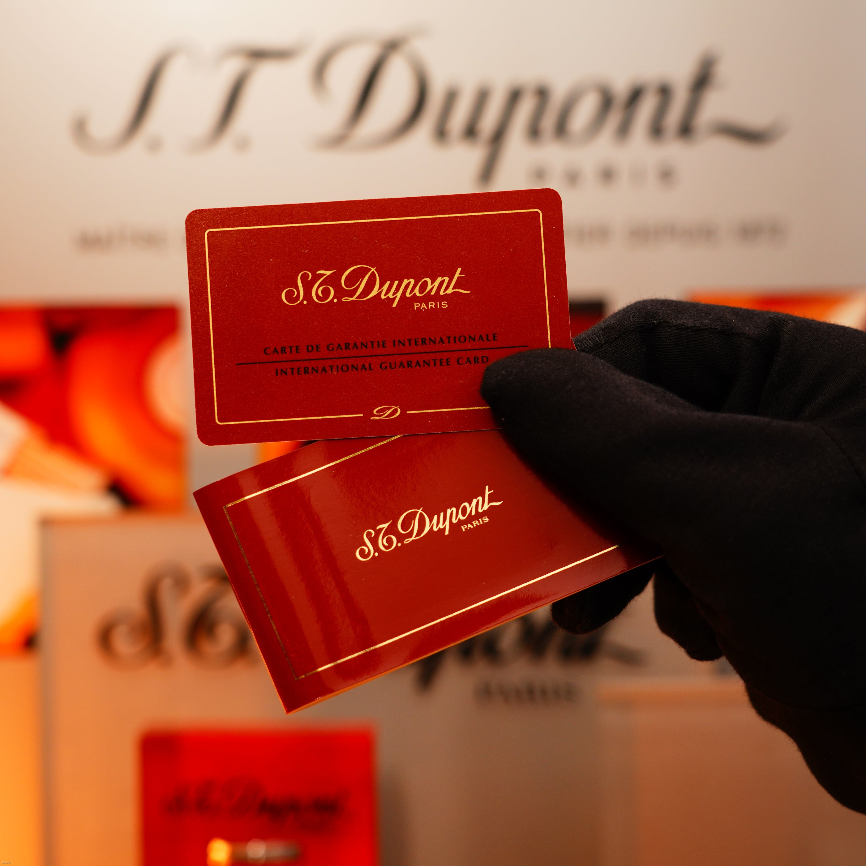A gloved hand holds two red Lucy Lighter guarantee cards, emphasizing the quality assurance, in front of a blurred background featuring the Vintage 1970 Extremely Rare 1/1 De St Dupont Solid 925 Sterling Ligne 1 Heavy Guilloché Pattern Lighter.