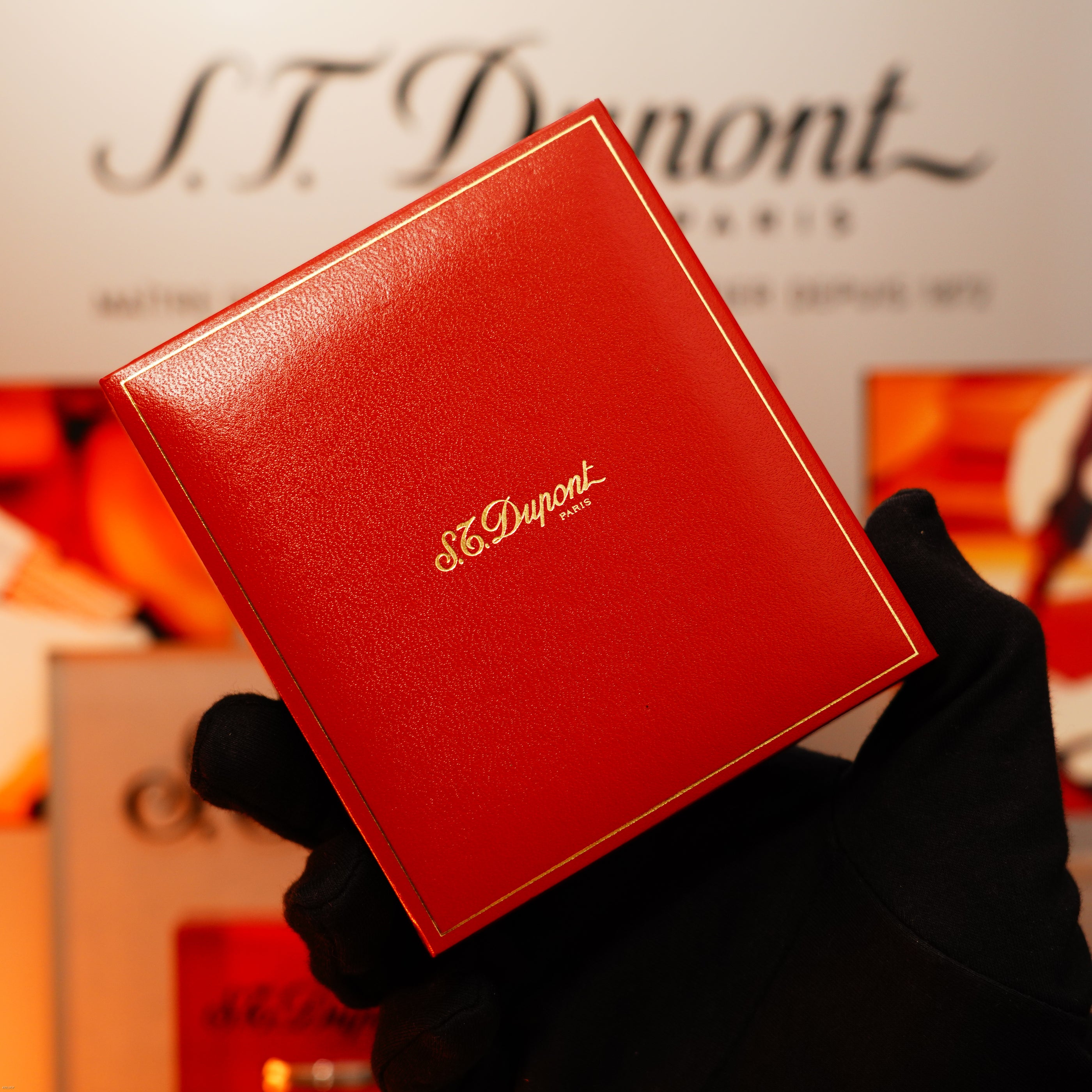 A gloved hand holds a red Lucy Lighter box with gold lettering, showcasing a Vintage 1970 Extremely Rare 1/1 De St Dupont Solid 925 Sterling Ligne 1 Heavy Guilloché Pattern Lighter, with a blurred Lucy Lighter sign in the background.
