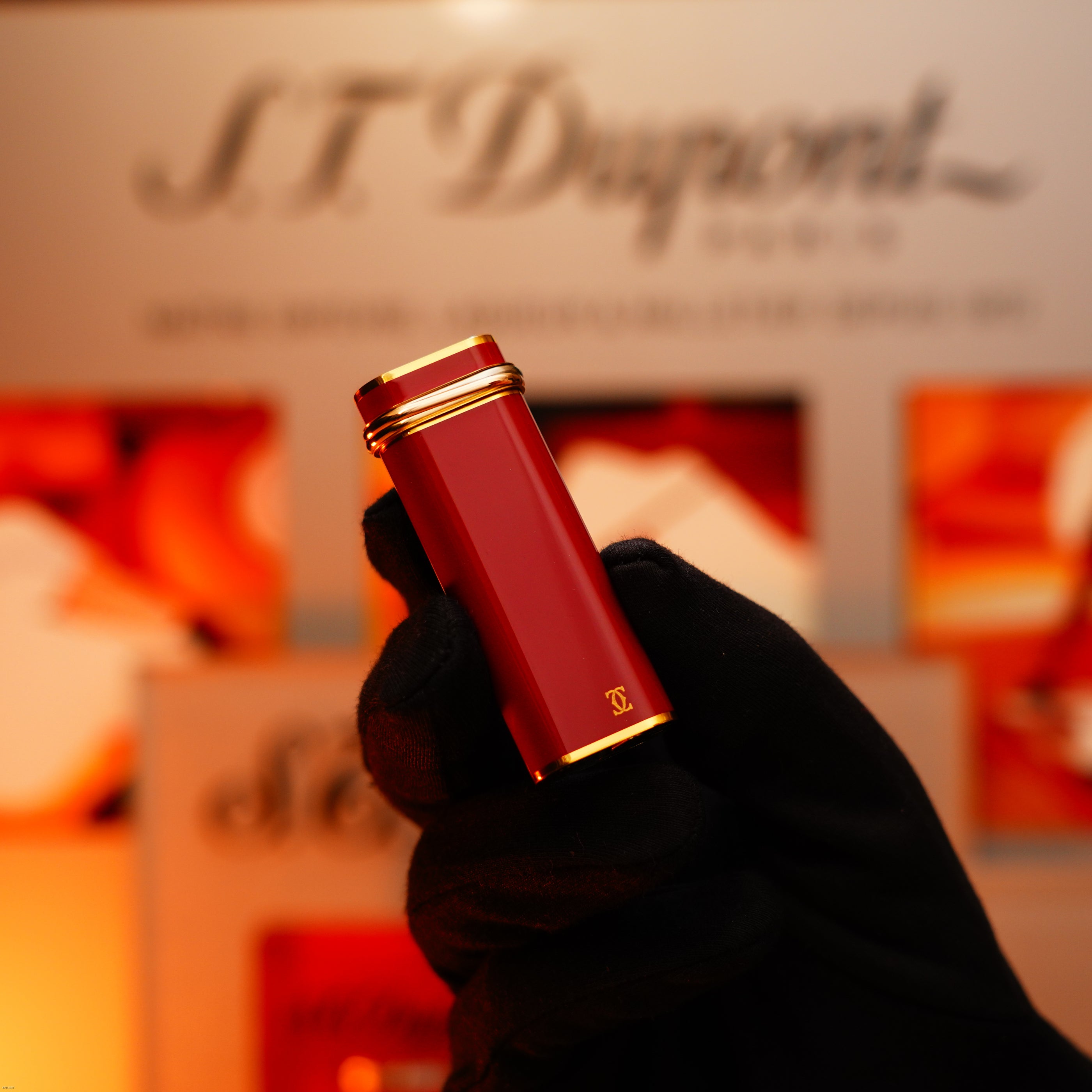 A gloved hand holds a Vintage 1980 Cartier Early Oval 24k Gold Finish Red Passion Lacquer Le Must Lighter by Lucy Lighter, with the brand name blurred in the background.