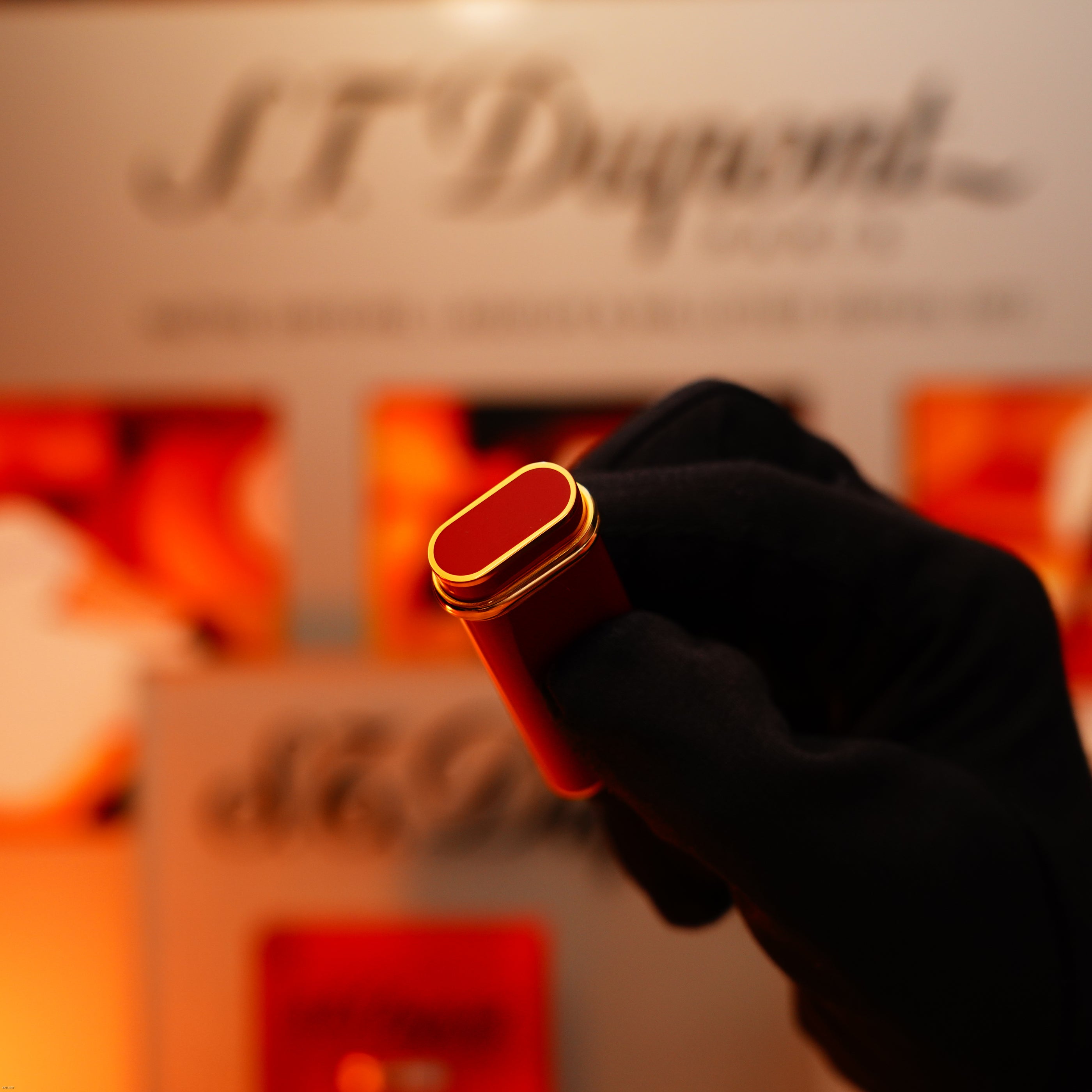A gloved hand holds a Vintage 1980 Cartier Early Oval 24k Gold Finish Red Passion Le Must Lighter by Lucy Lighter, featuring iconic Cartier lacquer and an 18k Trinity ring, in front of a blurred display.