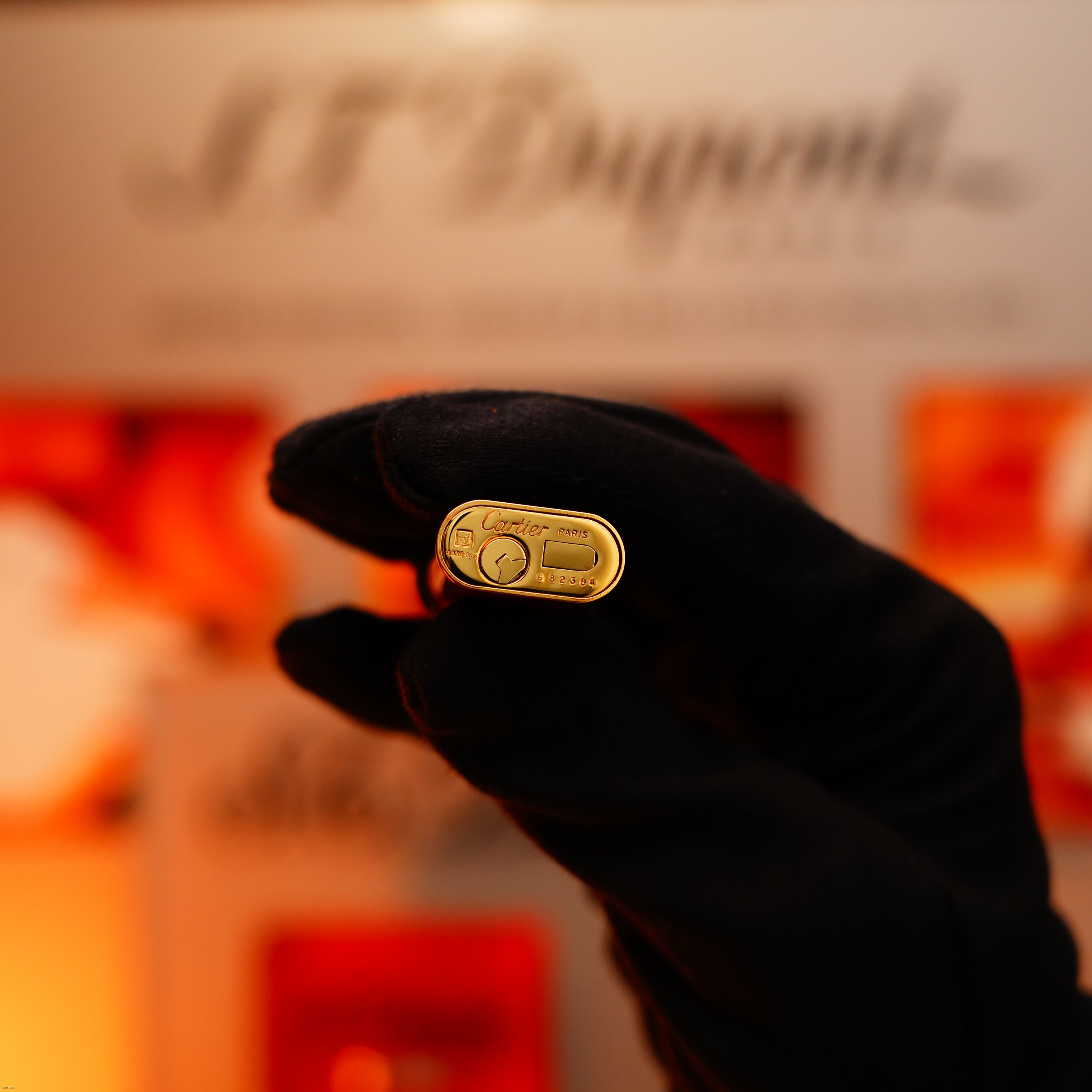 A gloved hand holds a rare Lucy Lighter vintage 1970 heavy silver finish guilloché pattern lighter, with a blurred ST Dupont sign in the background—showcasing classic design and craftsmanship.