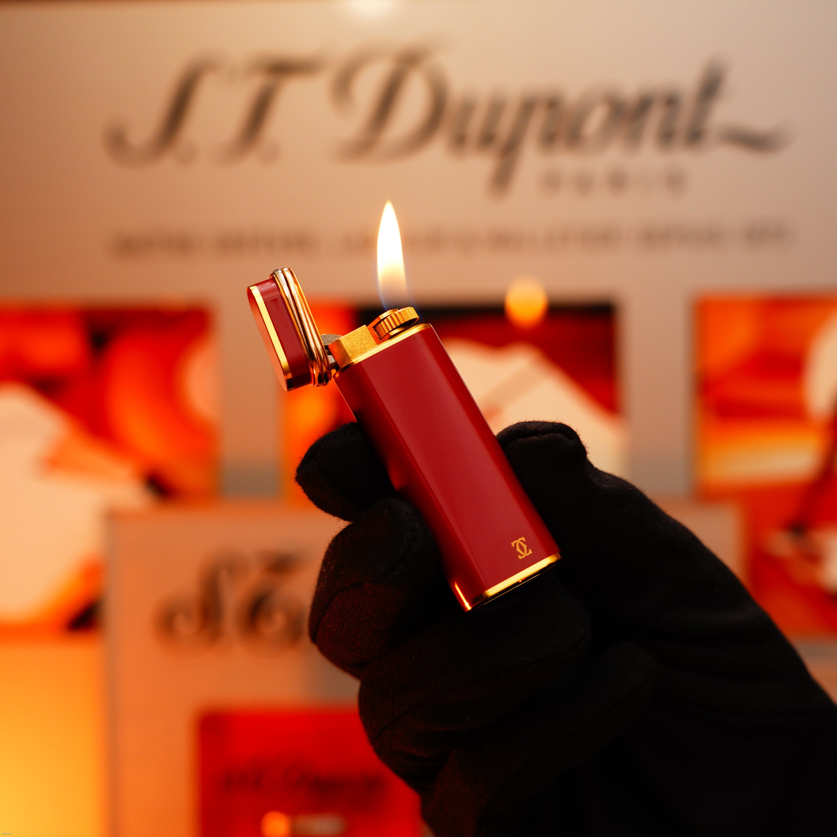 A gloved hand holds a Vintage 1980 Cartier Early Oval 24k Gold Finish Red Passion Le Must Lighter by Lucy Lighter with the lid open and flame lit, set against a branded background.