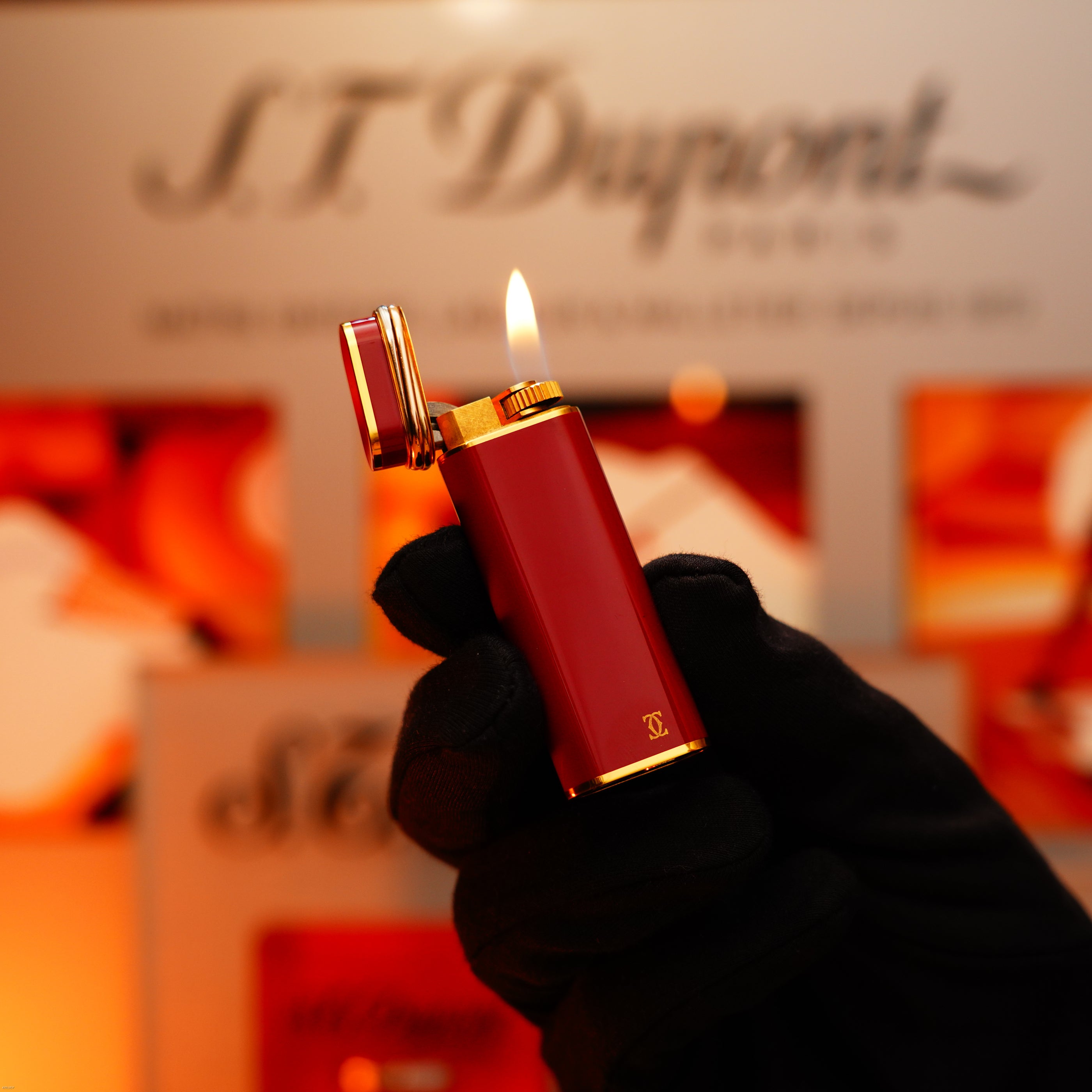 A gloved hand holds a Vintage 1980 Cartier Early Oval 24k Gold Finish Red Passion Le Must Lighter by Lucy Lighter, its lid open and flame ignited, against a blurred sign and warm background.