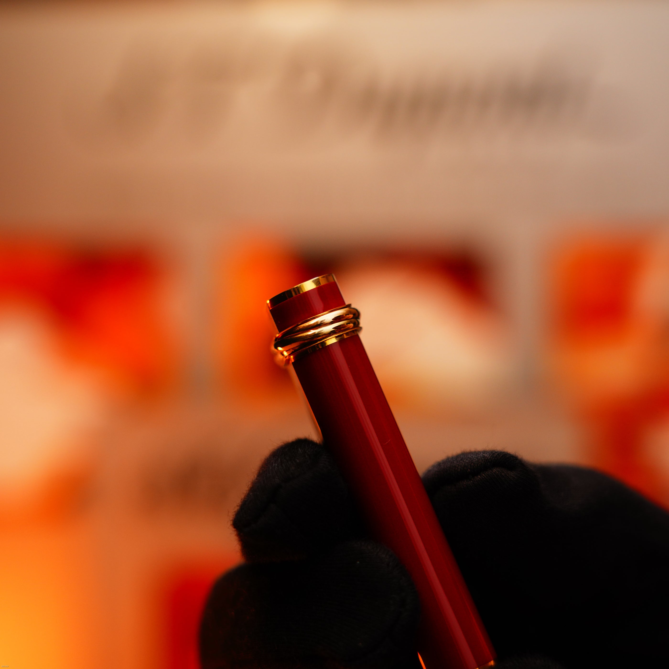 A gloved hand holds the Lucy Lighter Vintage 1980 Cartier Early Oval—red lacquer with a 24k gold finish and an 18k Trinity ring—its elegant design echoing vintage luxury, set against a softly blurred, warm background.