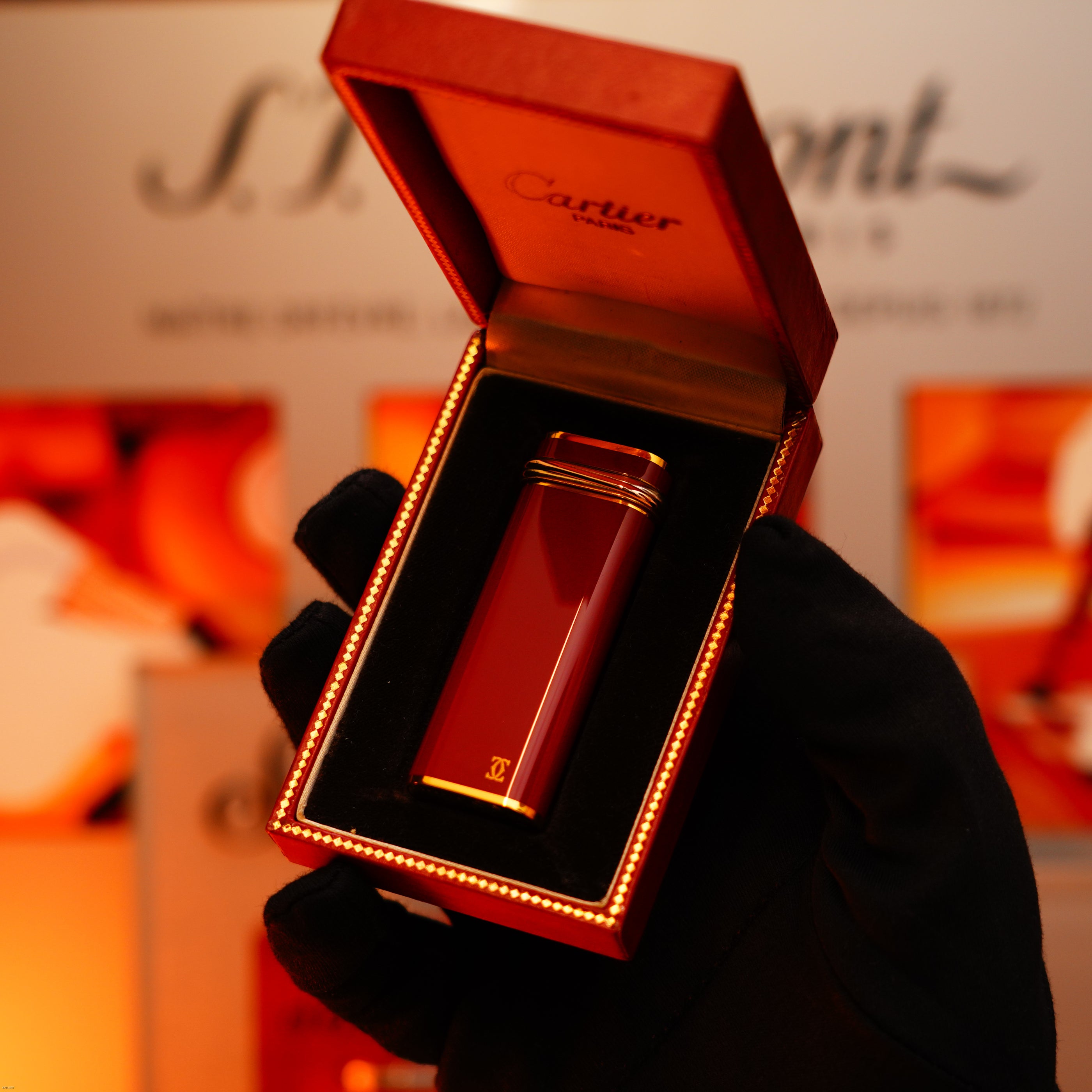 A gloved hand holds an open box displaying a Vintage 1980 Lucy Lighter Cartier Early Oval Red Passion Lacquer Le Must Lighter with 24k gold finish and 18k Trinity Ring, against a blurred background.