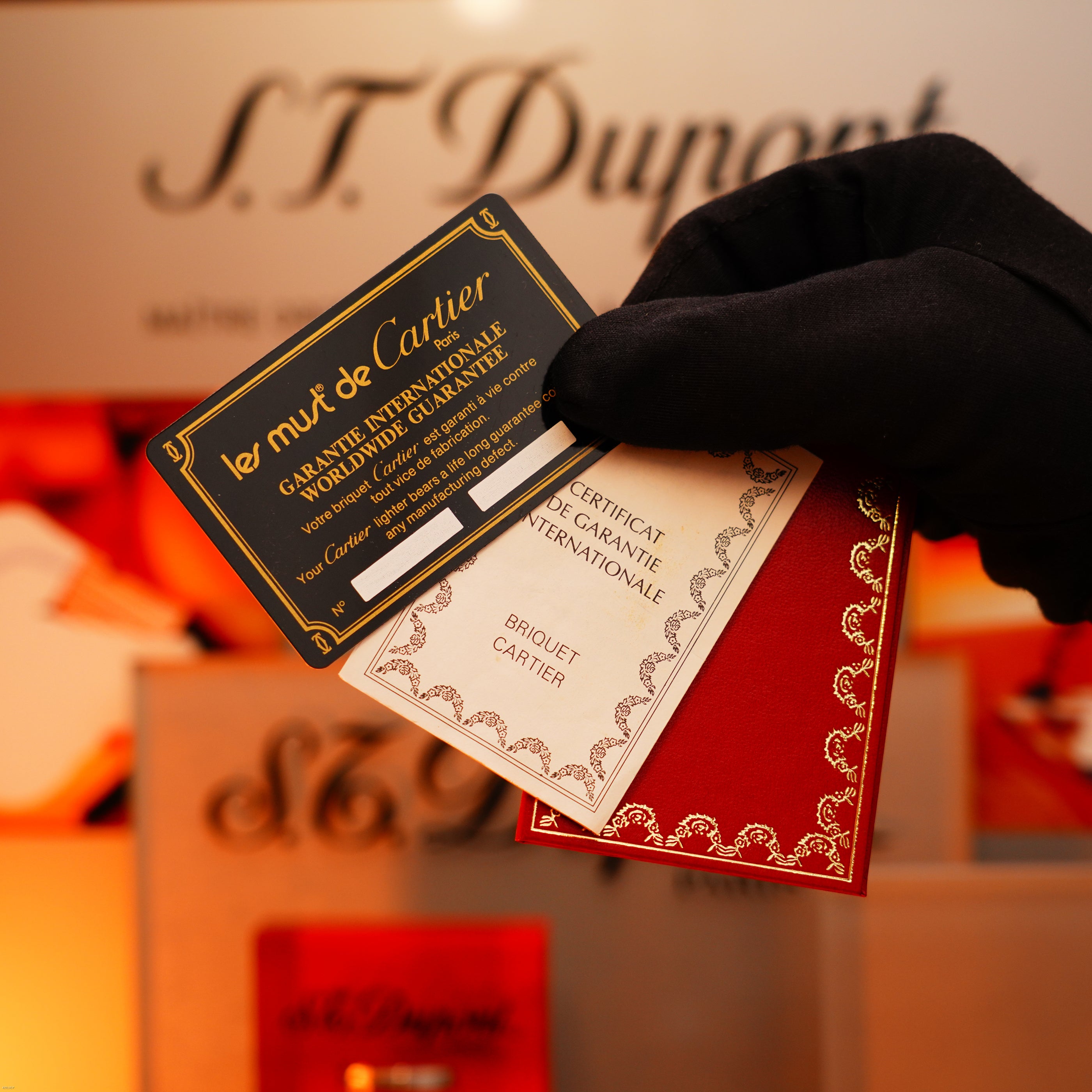 A gloved hand holds Cartier warranty cards and a certificate, attesting to quality, before Lucy Lighter packaging—celebrating the Vintage 1980 Cartier Early Oval 24k Gold Red Passion Lacquer 18k Trinity Ring Le Must Lighter.