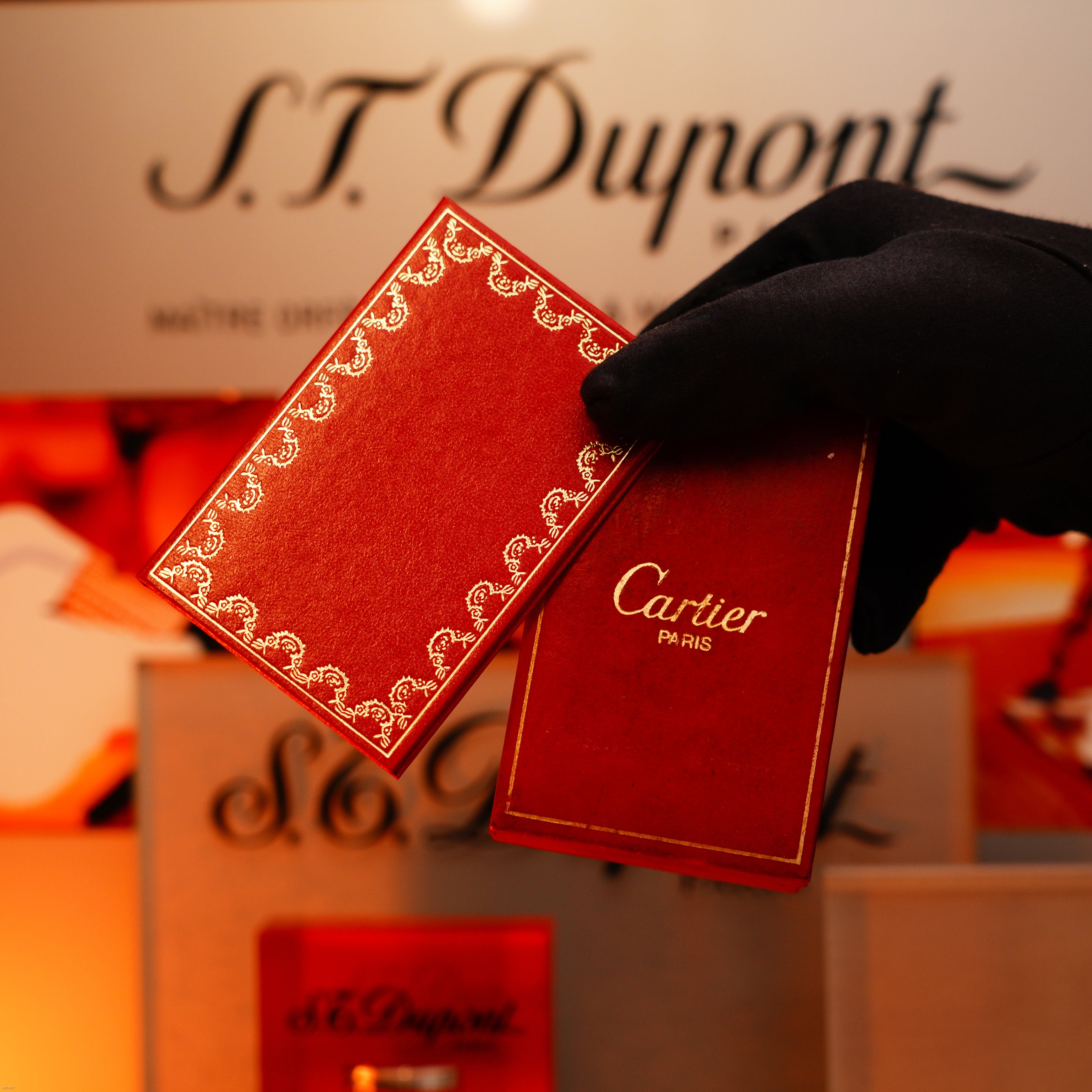 A gloved hand holds two luxury red cases, one reading “Cartier Paris,” in front of an S.T. Dupont sign—perfect for displaying the Lucy Lighter Vintage 1980 Cartier Early Oval 24k Gold Finish Le Must Lighter with 18k Trinity Ring.