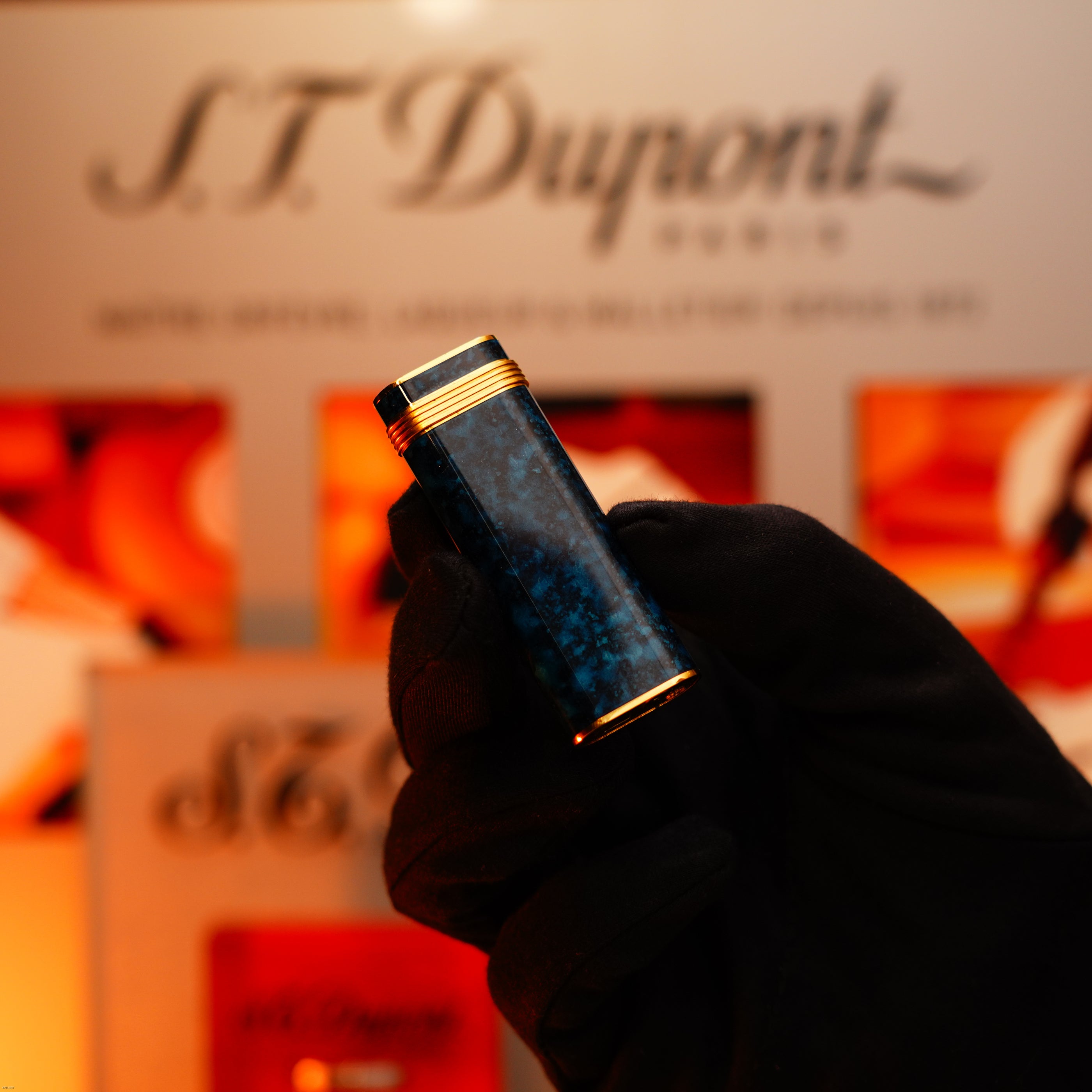 A gloved hand holds a Vintage 1980 Cartier Oval Le Must Lighter by Lucy Lighter, featuring a 24k gold finish, lapiz lacquer body, and an 18k Trinity ring on the door against a blurred display background.