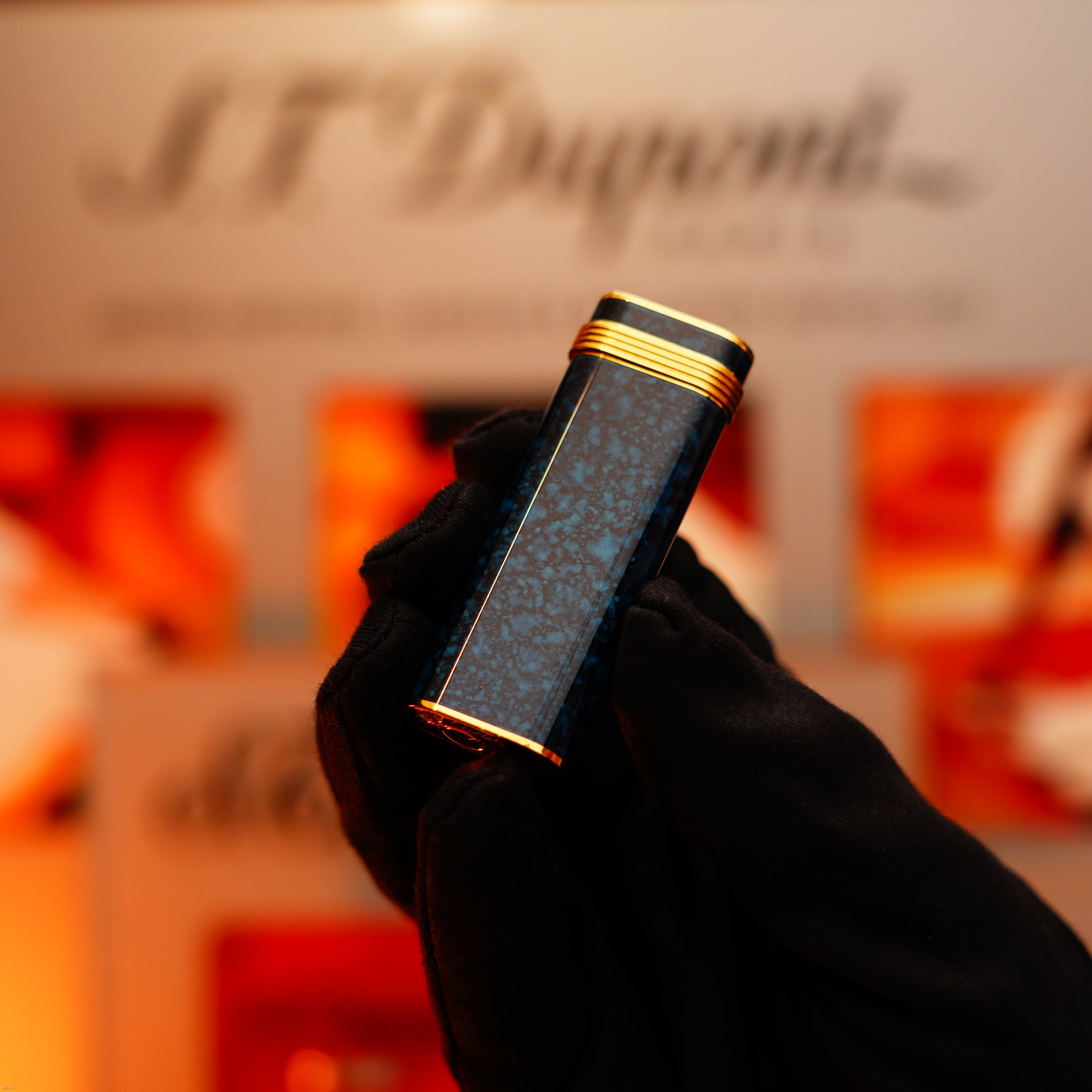 A gloved hand holds a Vintage 1980 Cartier Oval Lucy Lighter, 24k gold finish with lapiz lacquer and an 18k Trinity ring on the door, in front of a blurred sign—highlighting its timeless luxury craftsmanship.