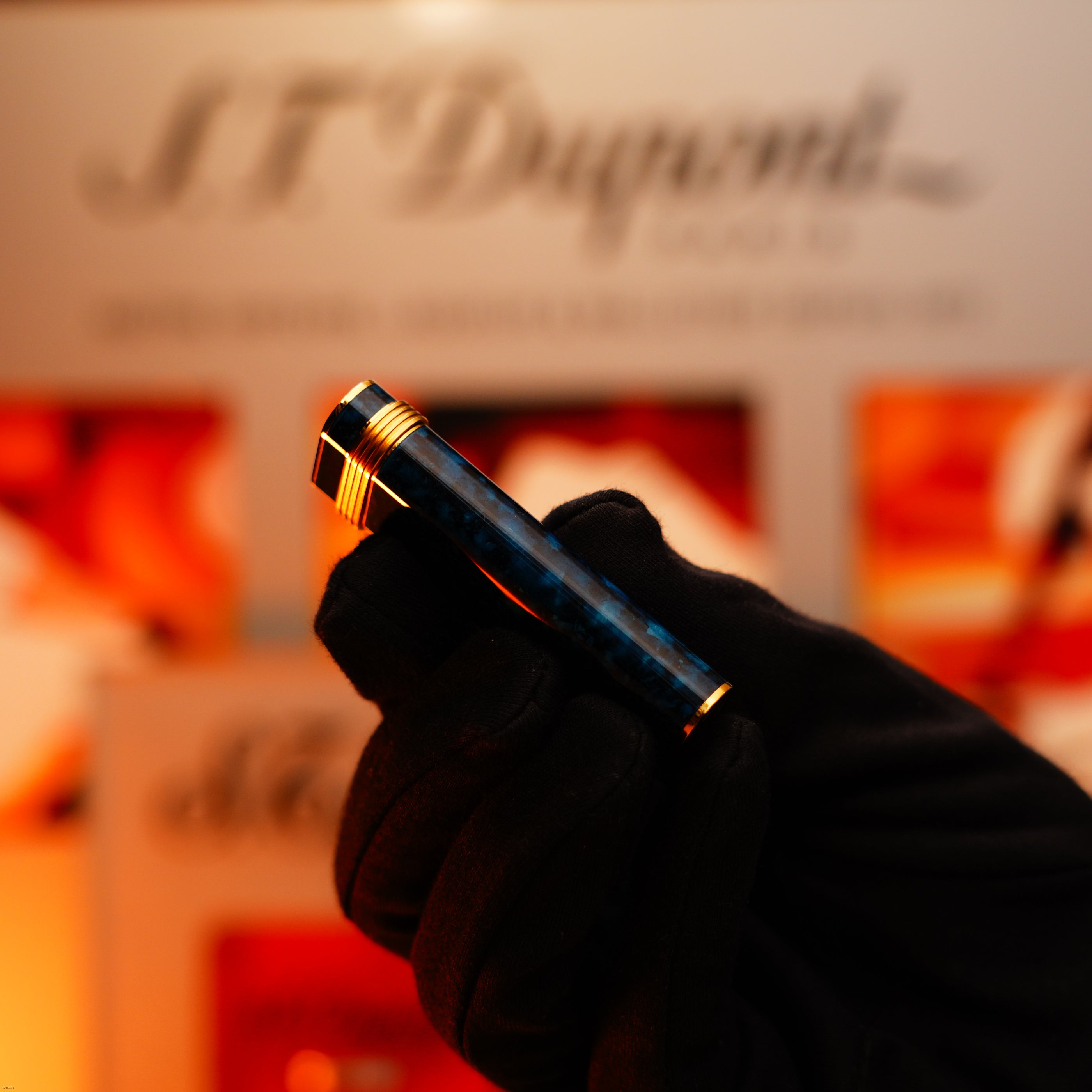 A gloved hand holds the Vintage 1980 Cartier Oval 24k Gold Finish Lapiz Lacquer with 18k Trinity Ring On Door Le Must Lighter by Lucy Lighter, its luxury design highlighted against a warm, blurred backdrop—an emblem of timeless quality.