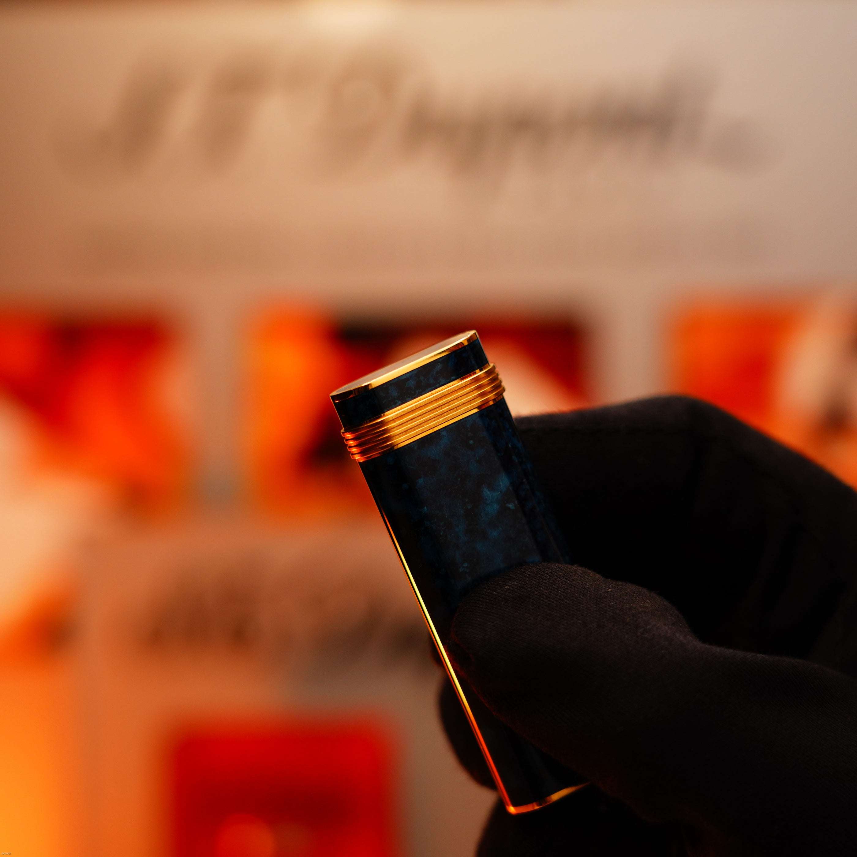 A gloved hand holds a Lucy Lighter: Vintage 1980 Cartier Oval 24k Gold Finish Lapiz Lacquer with 18k Trinity Ring on Door Le Must Lighter, set against a blurred background with warm lighting.