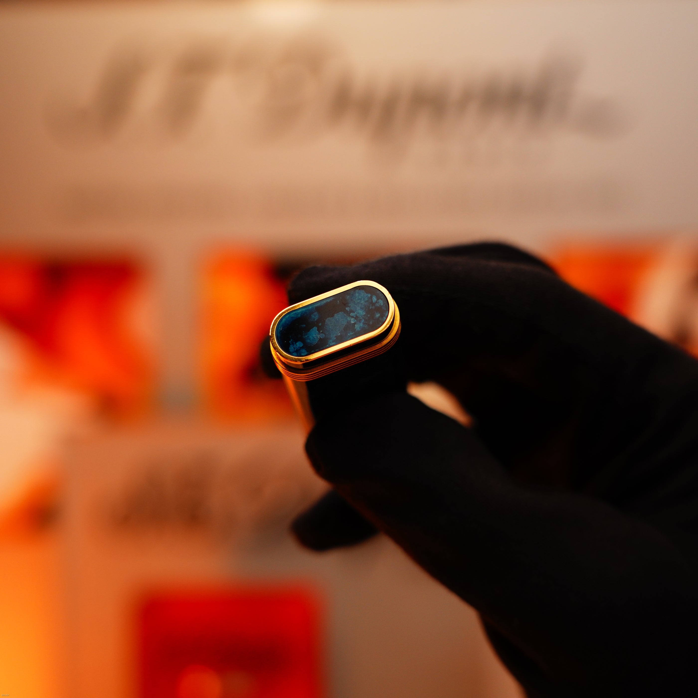 A gloved hand holds a Vintage 1980 Cartier Oval 24k Gold Finish Lapiz Lacquer Lighter with 18k Trinity Ring by Lucy Lighter, in front of a blurred display background.