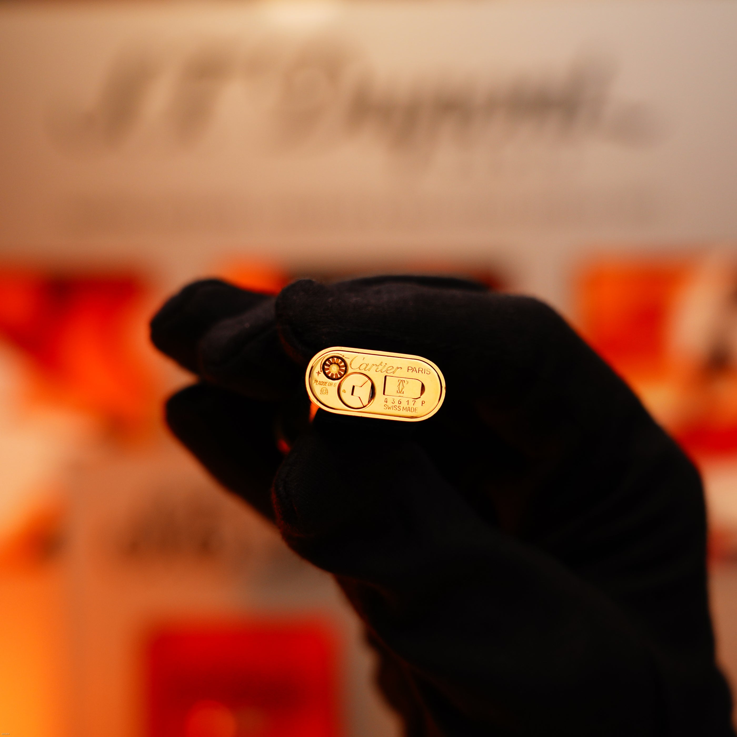 A gloved hand holds a Vintage 1980 Cartier Oval 24k Gold Finish Lapiz Lacquer Le Must Lighter with an 18k Trinity Ring on the door by Lucy Lighter, against a blurred background.