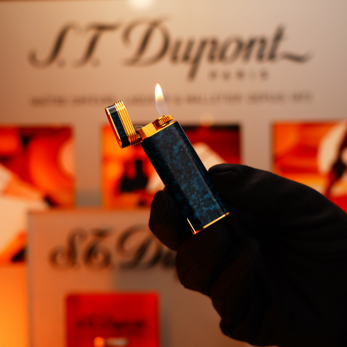 A gloved hand holds a Lucy Lighter Vintage 1980 Cartier Oval Le Must Lighter, finished in 24k gold with lapiz lacquer and an 18k Trinity ring on the door, flames flickering before a blurred display.