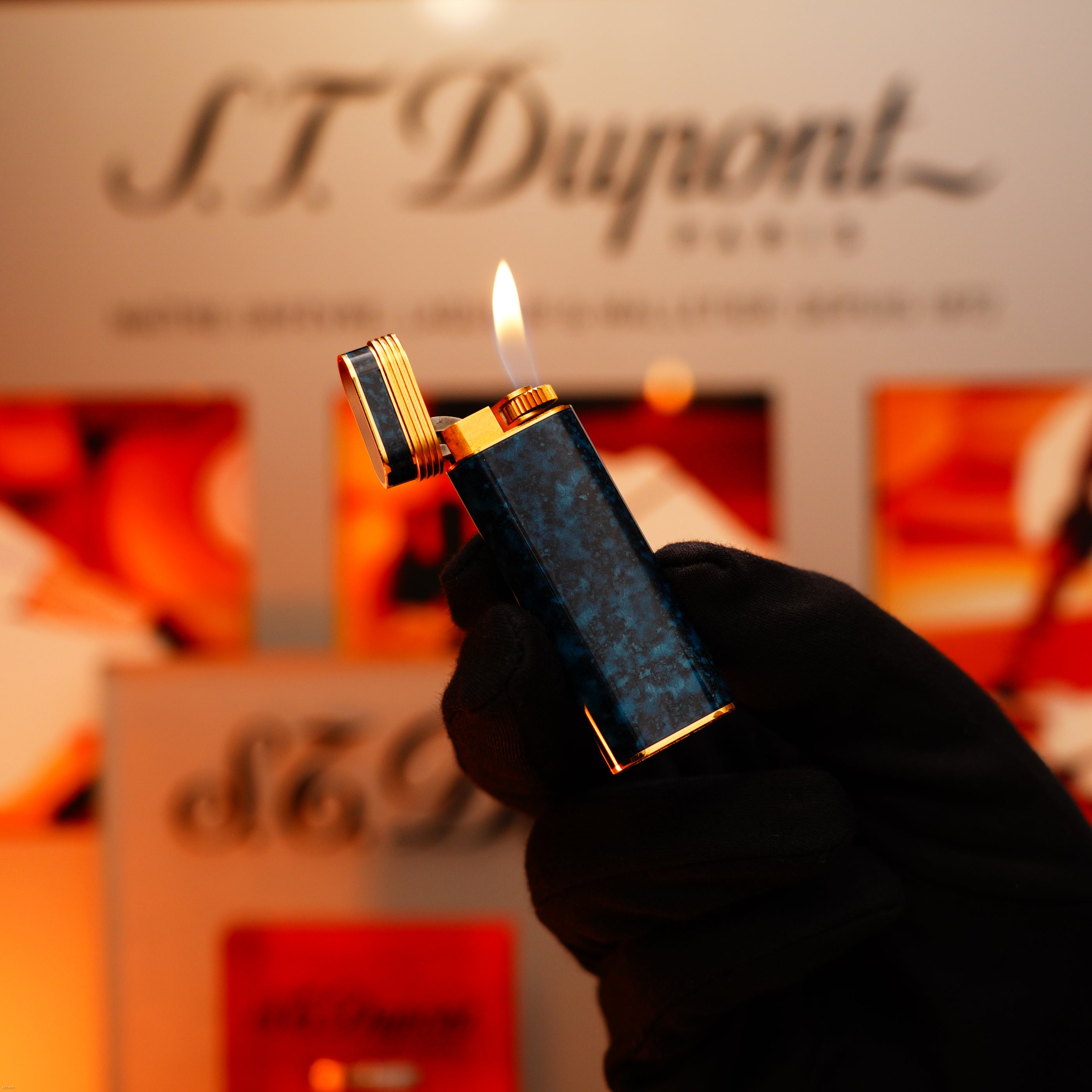 A gloved hand holds a Vintage 1980 Cartier Oval 24k Gold Finish Lapiz Lacquer lighter with an 18k Trinity ring on the door by Lucy Lighter, its lid open and flame ignited against a blurred branded background.