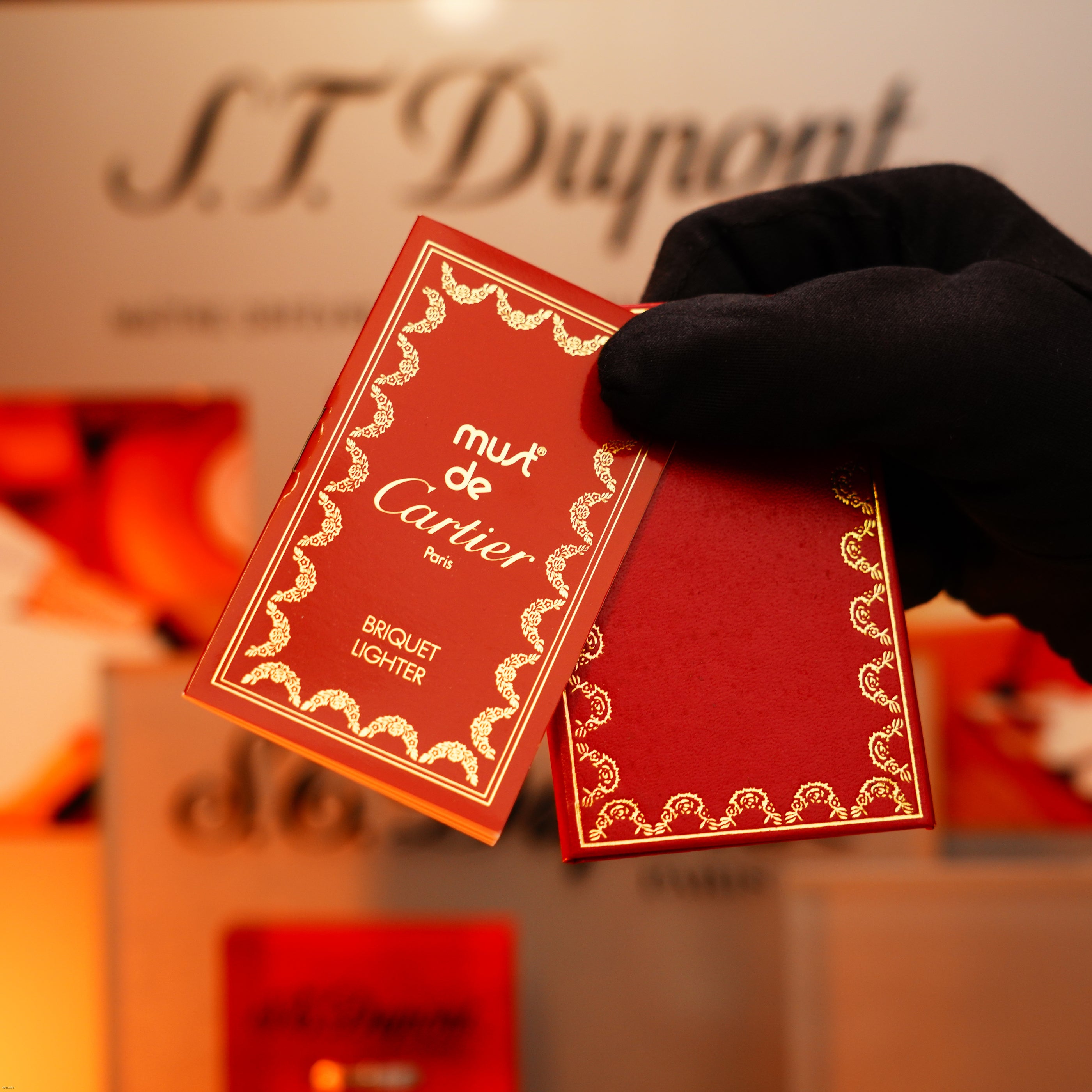 A gloved hand holds a Lucy Lighter Vintage 1980 Cartier Oval 24k Gold Finish Lapiz Lacquer lighter with an 18k Trinity Ring on the door, showcased against a blurred ST Dupont sign in the background.