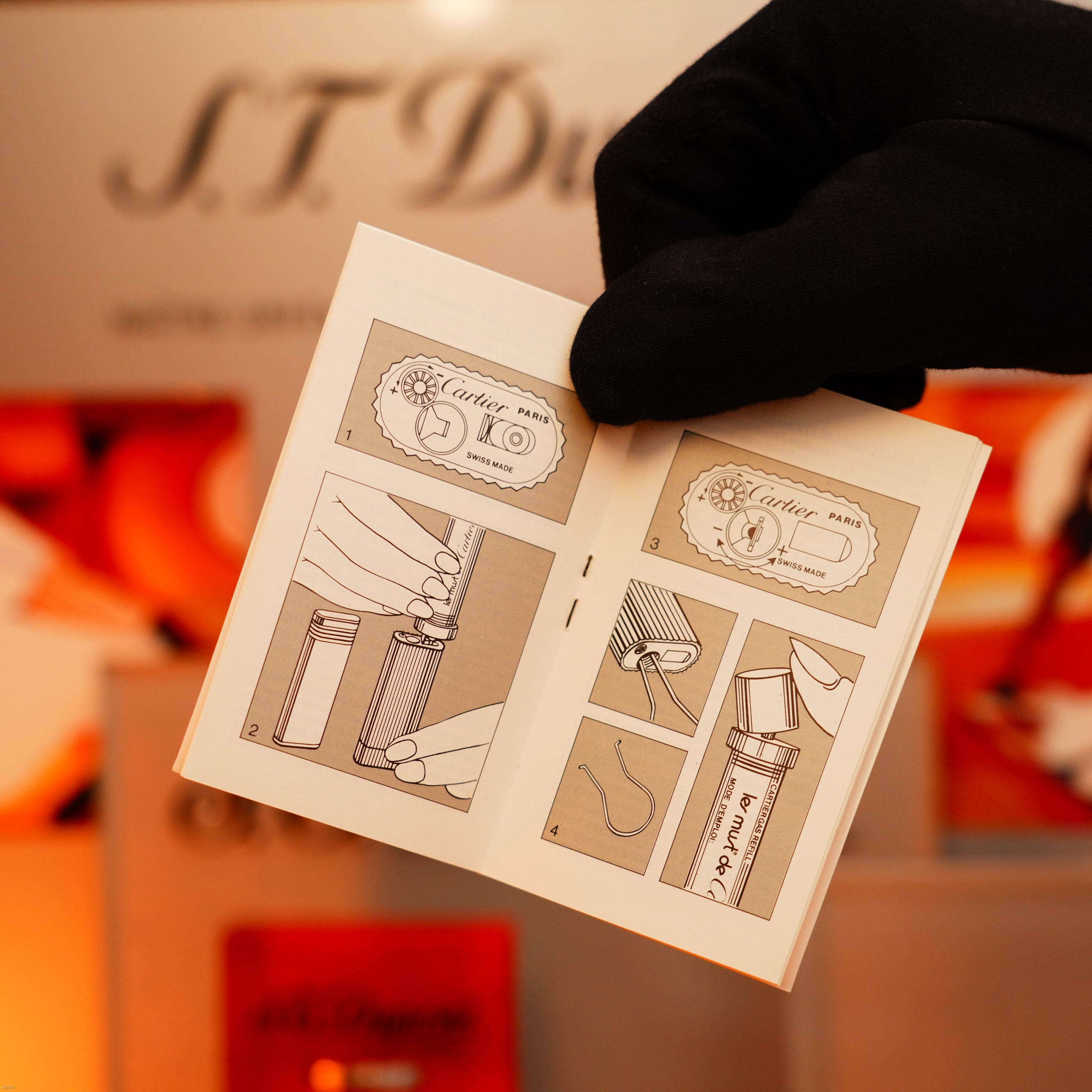 A gloved hand holds an open instruction booklet with illustrated steps for using the Lucy Lighter Vintage 1980 Cartier Oval 24k Gold Finish Lapiz Lacquer Le Must Lighter; blurred packaging appears in the background.