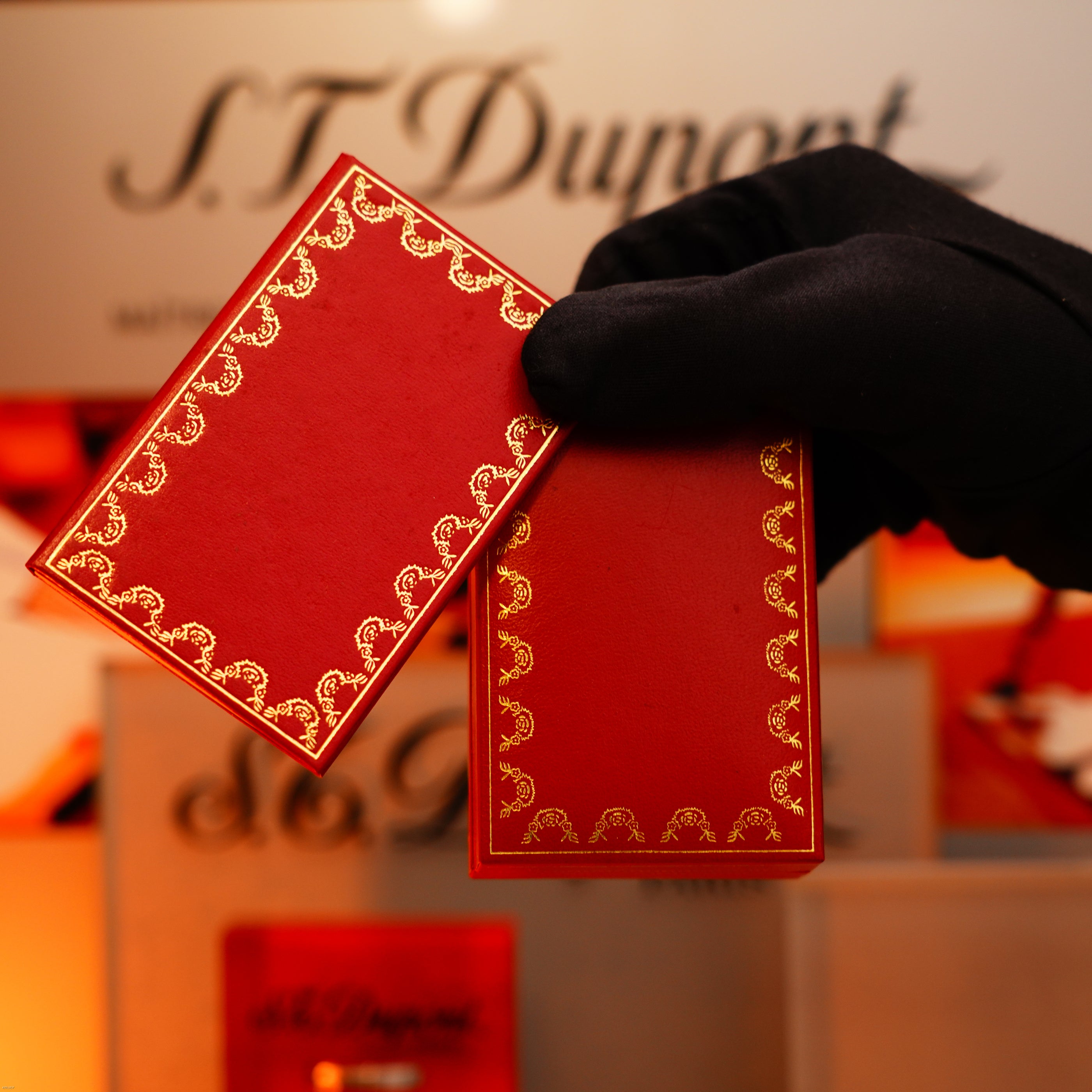 A gloved hand holds two red cards with gold borders in front of a Lucy Lighter display featuring the Vintage 1980 Cartier Oval 24k Gold Finish Lapiz Lacquer with 18k Trinity Ring Le Must Lighter, reflecting timeless craftsmanship.