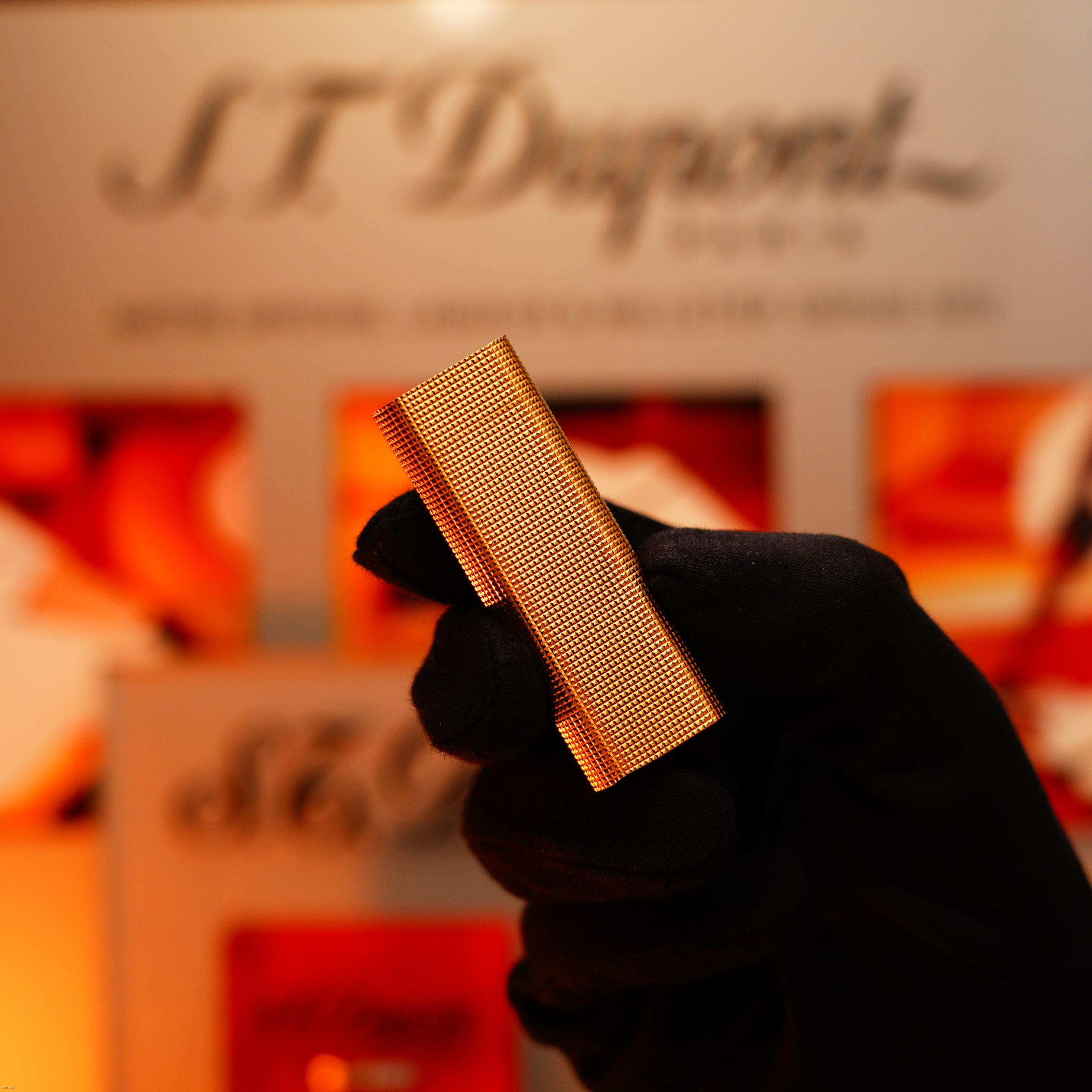 A gloved hand holds a Vintage 1970 Early Cartier Production Heavy Silver Finish Rare Guilloché Pattern Lighter by Lucy Lighter, set against a blurred branded display background.