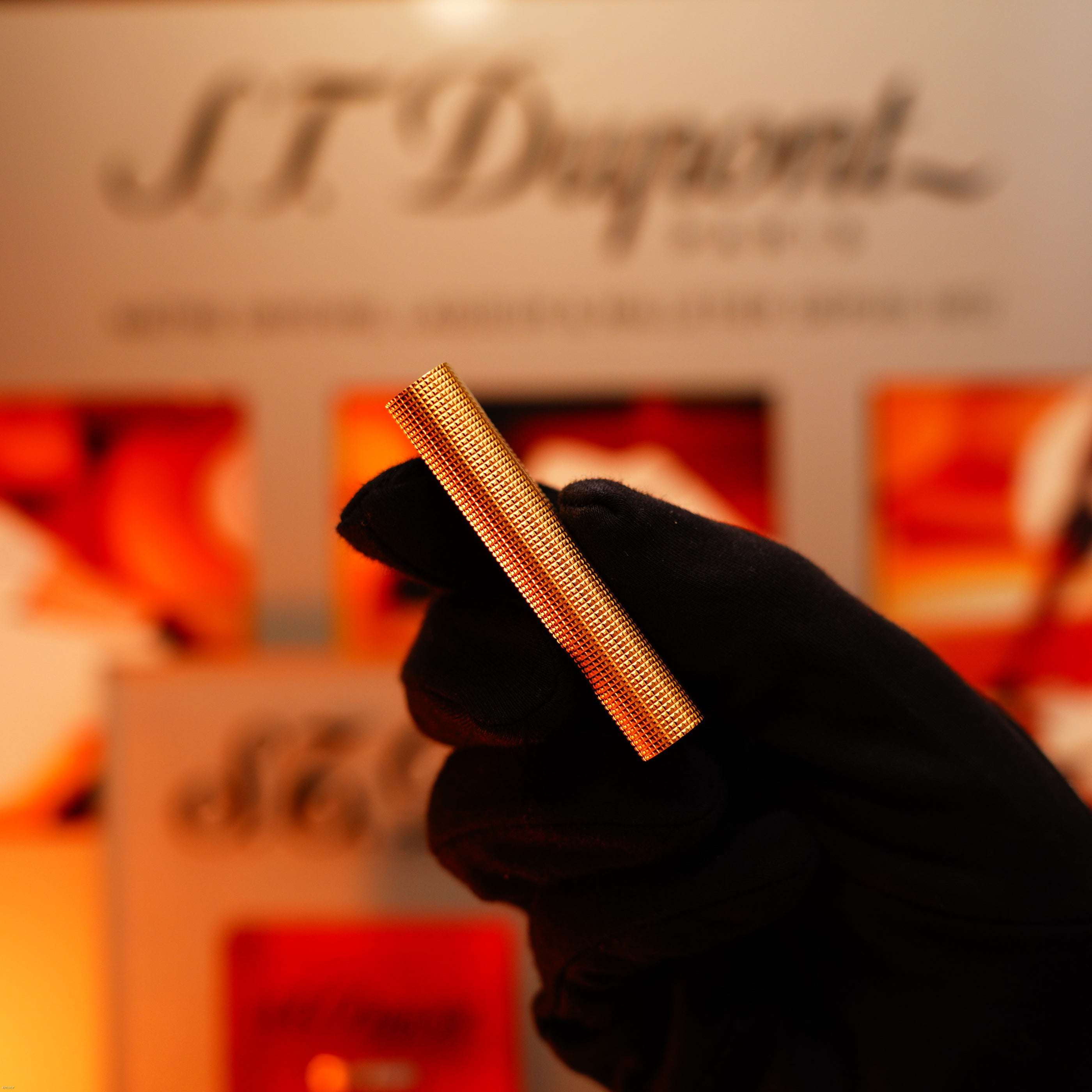 A gloved hand holds a Vintage 1970 Early Cartier Production Heavy Silver Finish Rare Guilloché Pattern Lighter by Lucy Lighter, with a blurred S.T. Dupont sign and orange-red objects in the background.