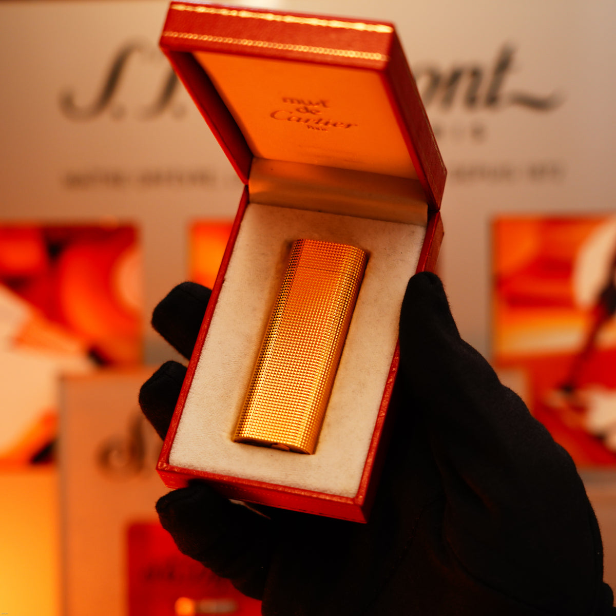 A gloved hand holds an open red box with a Vintage 1970 Early Cartier Production Heavy Silver Finish Rare Guilloché Pattern Lighter by Lucy Lighter, its surface gleaming against a blurred background with text and orange hues.