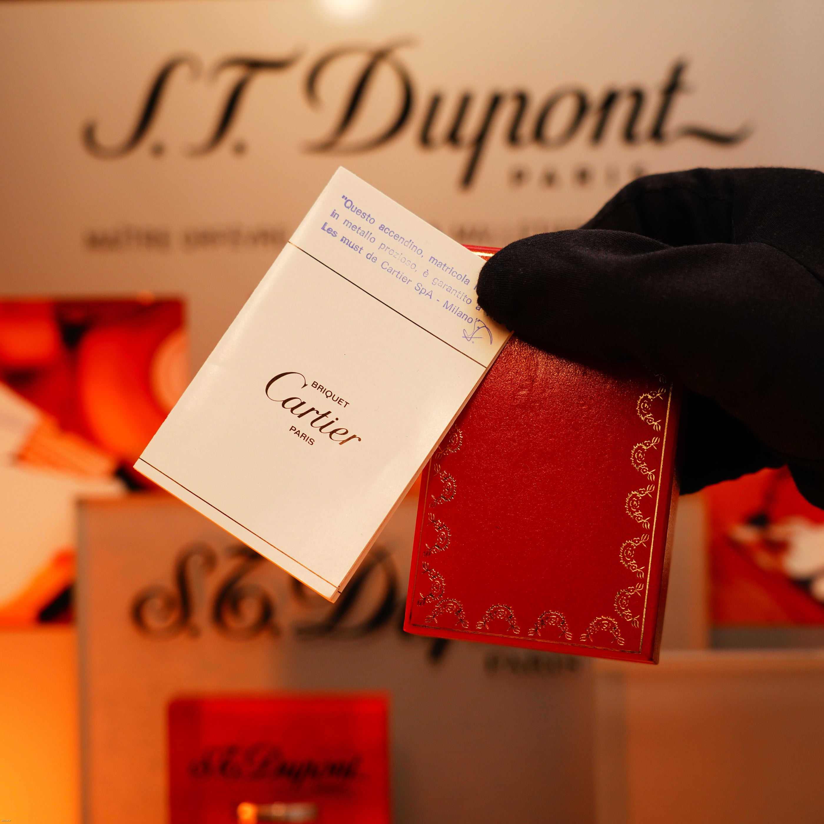 A gloved hand holds a Lucy Lighter Vintage 1970 Early Cartier Production Heavy Silver Finish Rare Guilloché Pattern Lighter in front of an S.T. Dupont Paris display, showcasing vintage luxury craftsmanship and repair services.