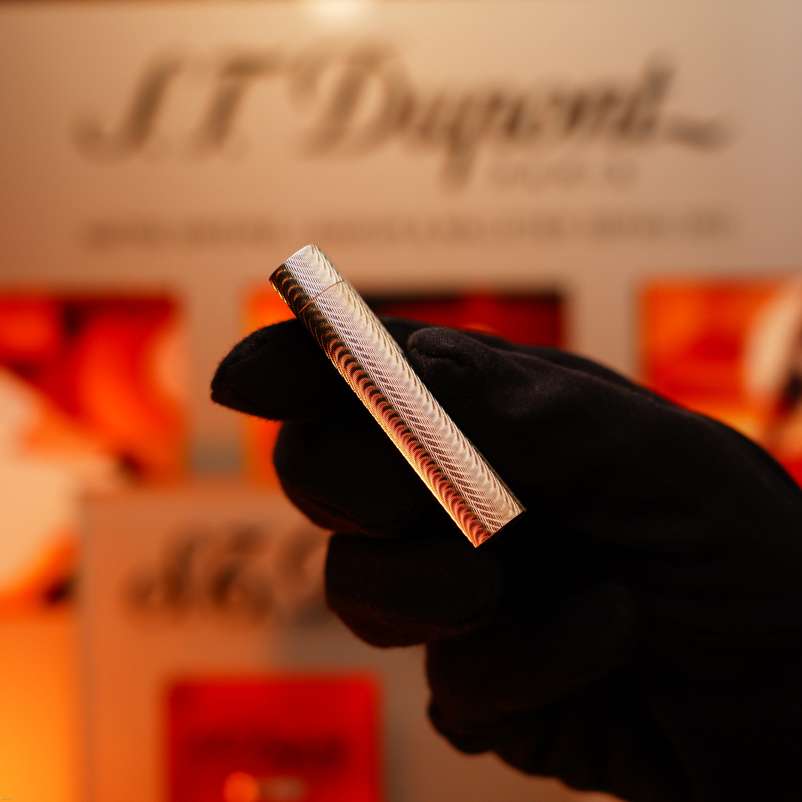 A gloved hand holds a Vintage 1970 Early Cartier Production Heavy Silver Rare Guilloché Pattern Lighter by Lucy Lighter in front of a blurred display, highlighting its metallic texture and luxury appeal.