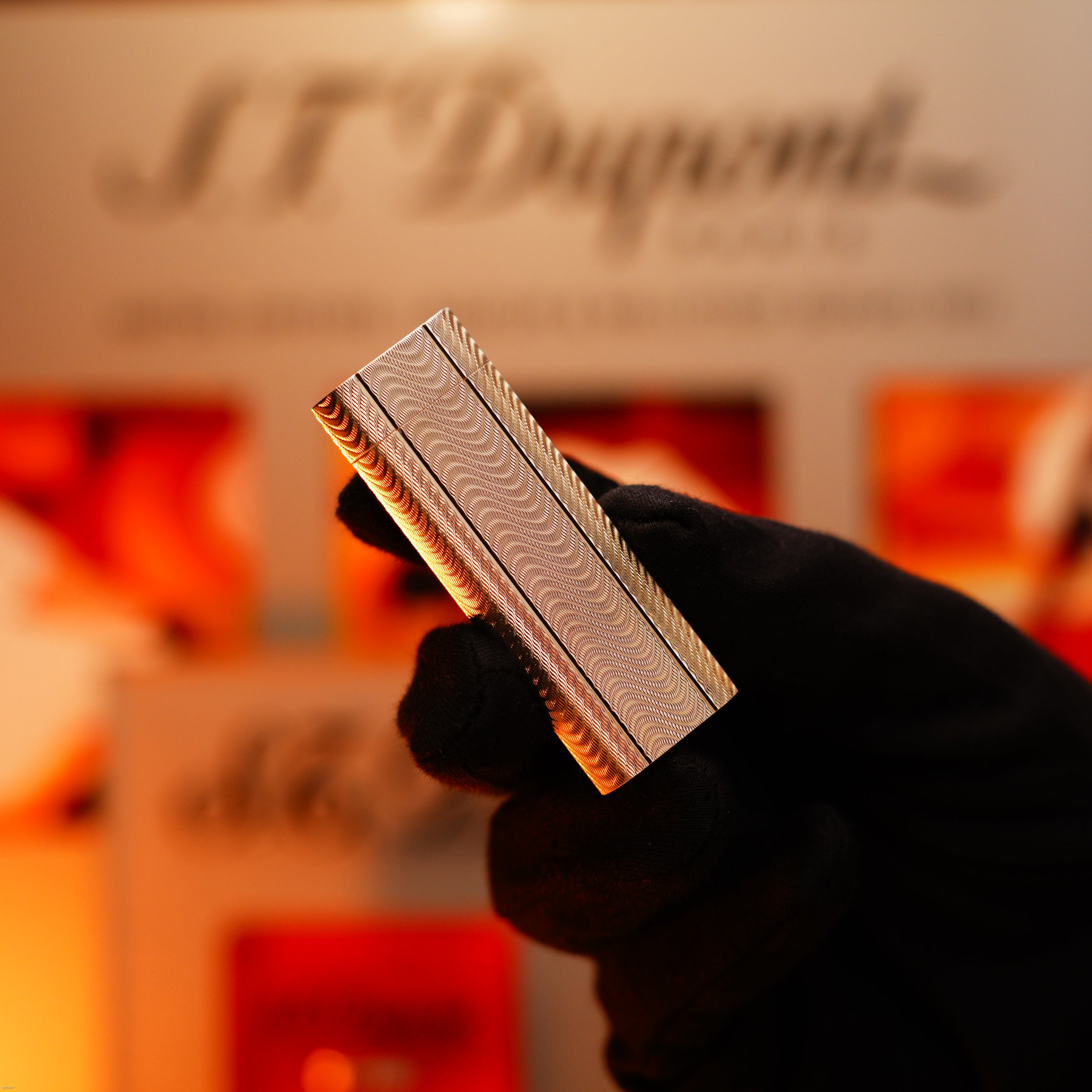 A gloved hand holds a Vintage 1970 Early Cartier Production Heavy Silver Rare Guilloché Pattern Lighter by Lucy Lighter—rectangular, silver, with a wavy pattern—against a blurred background showing the brand logo.