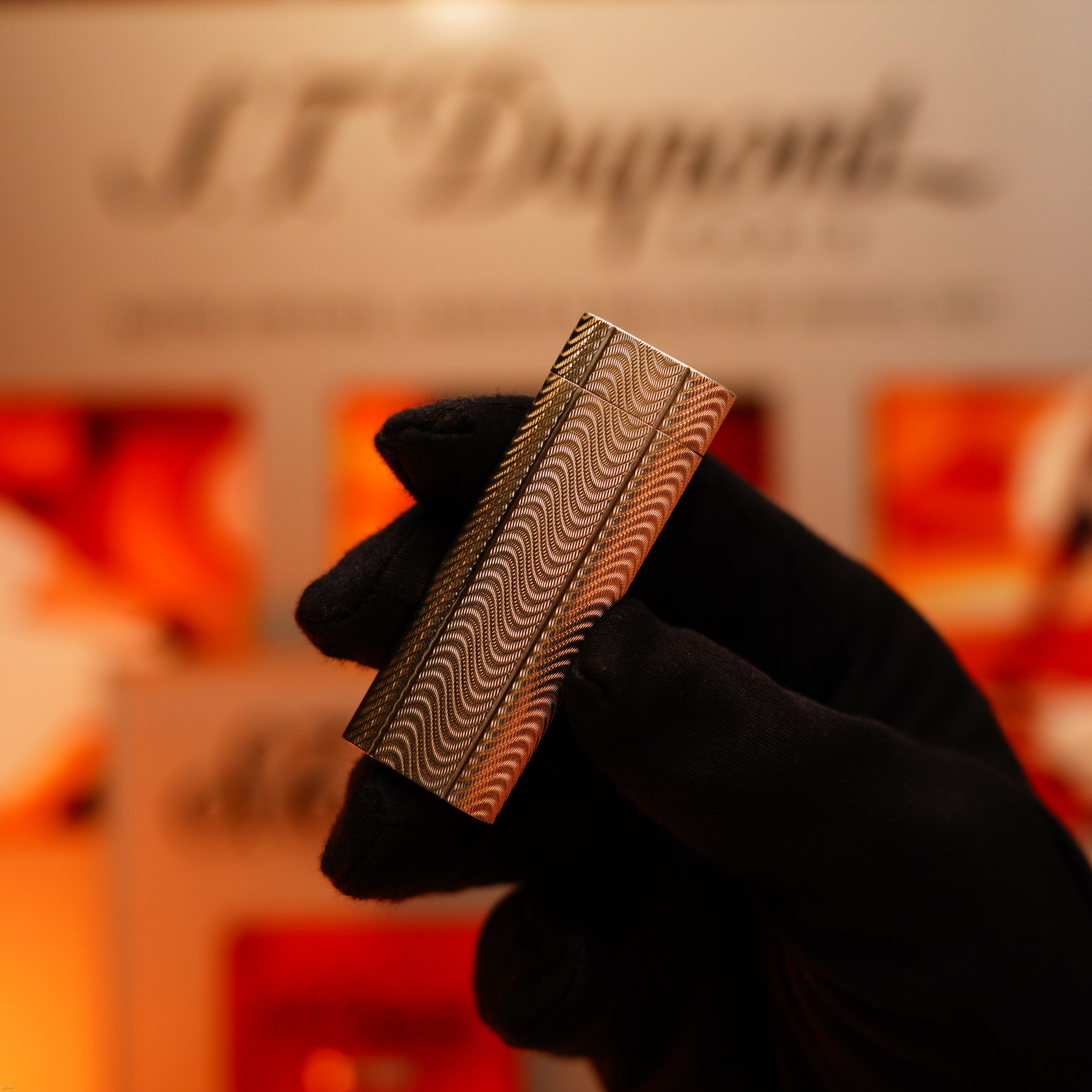 A gloved hand holds a heavy silver Vintage 1970 Early Cartier Production Lucy Lighter with a rare guilloché pattern, while the brand name appears blurred in the background.