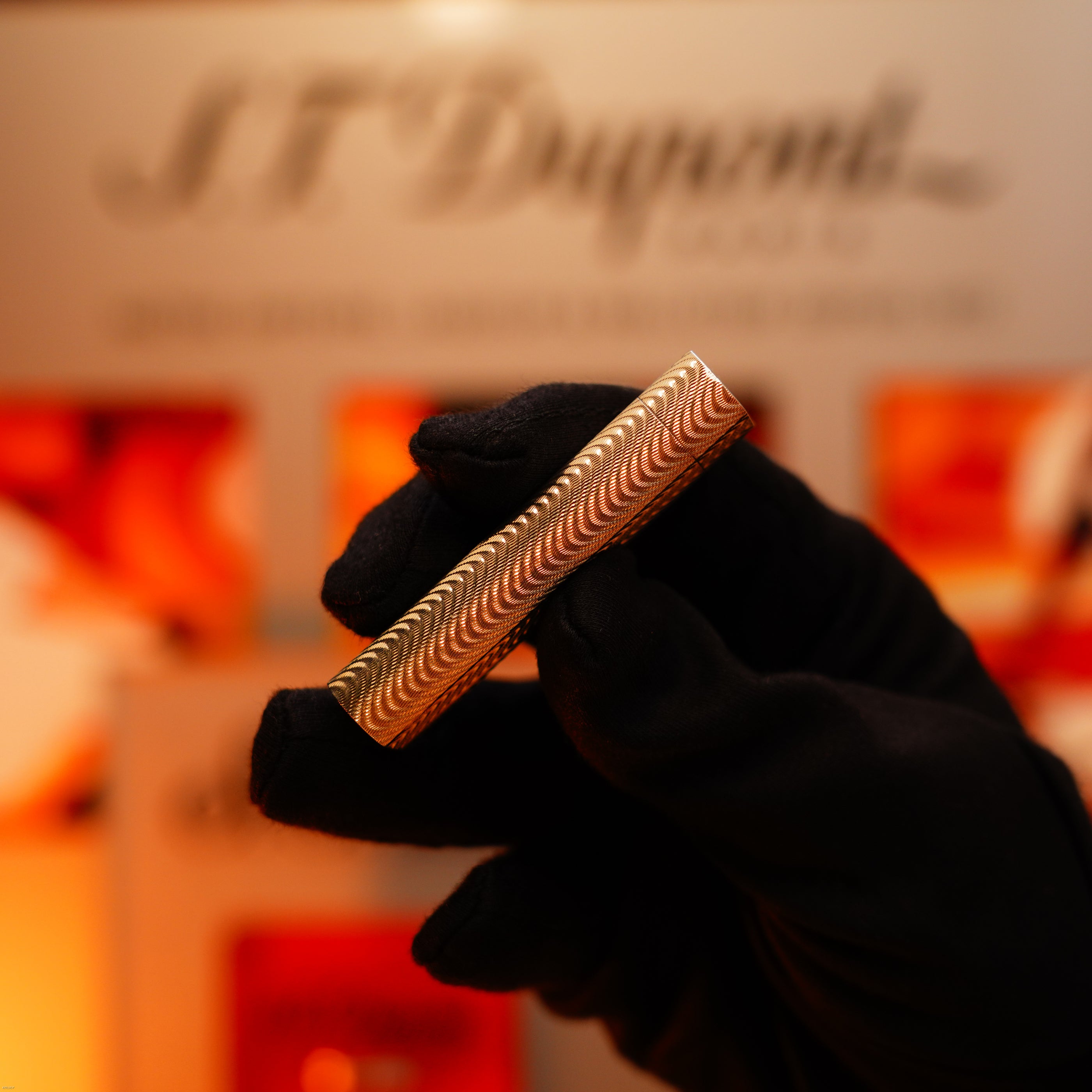 A gloved hand holds a Vintage 1970 Early Cartier Production Heavy Silver Rare Guilloché Pattern Lighter by Lucy Lighter, in front of a blurred S.T. Dupont Paris display.
