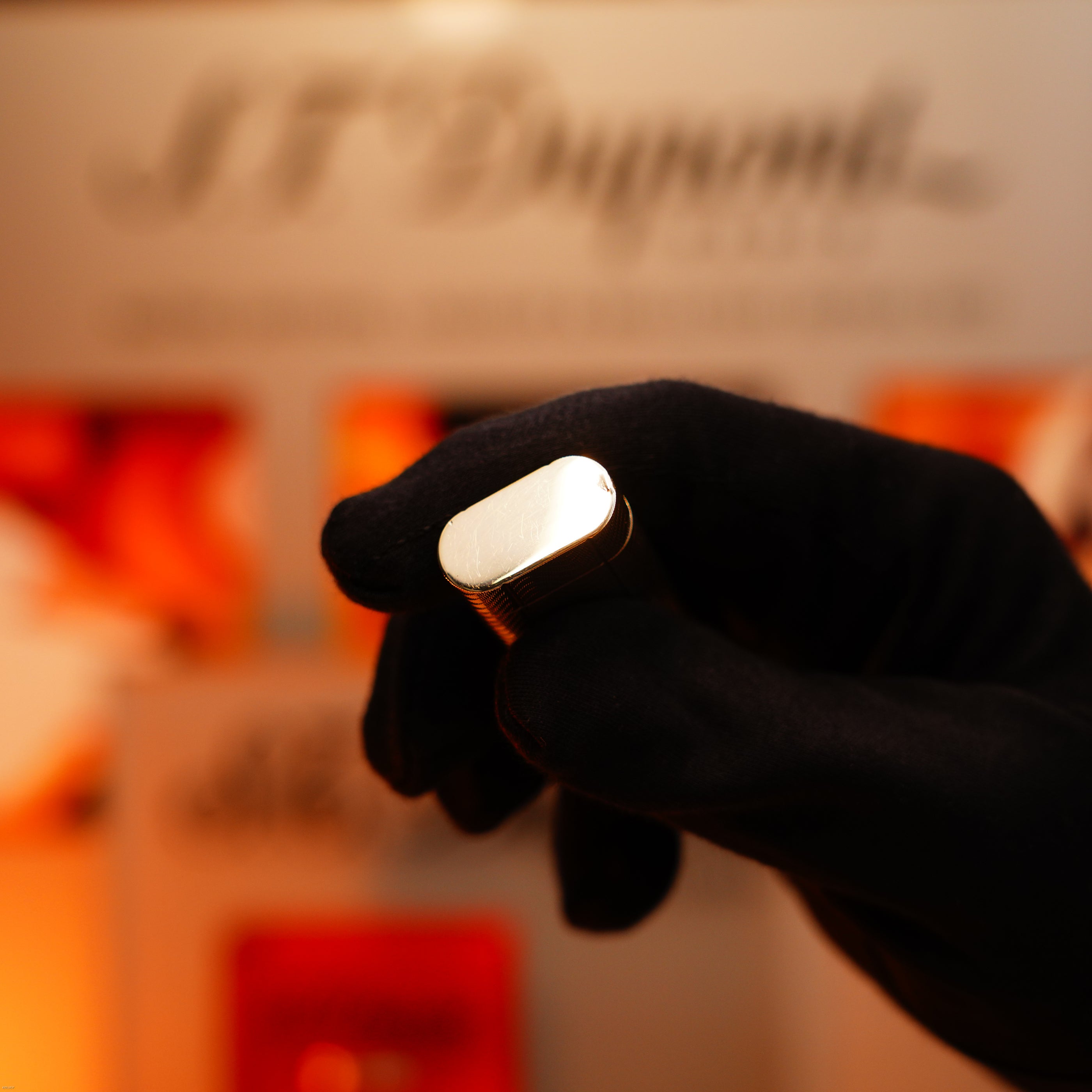 A gloved hand holds a Vintage 1970 Early Cartier Production Heavy Silver Rare Guilloché Pattern Lighter by Lucy Lighter, with a blurred background featuring the brand’s logo and assurance of quality and working warranty.