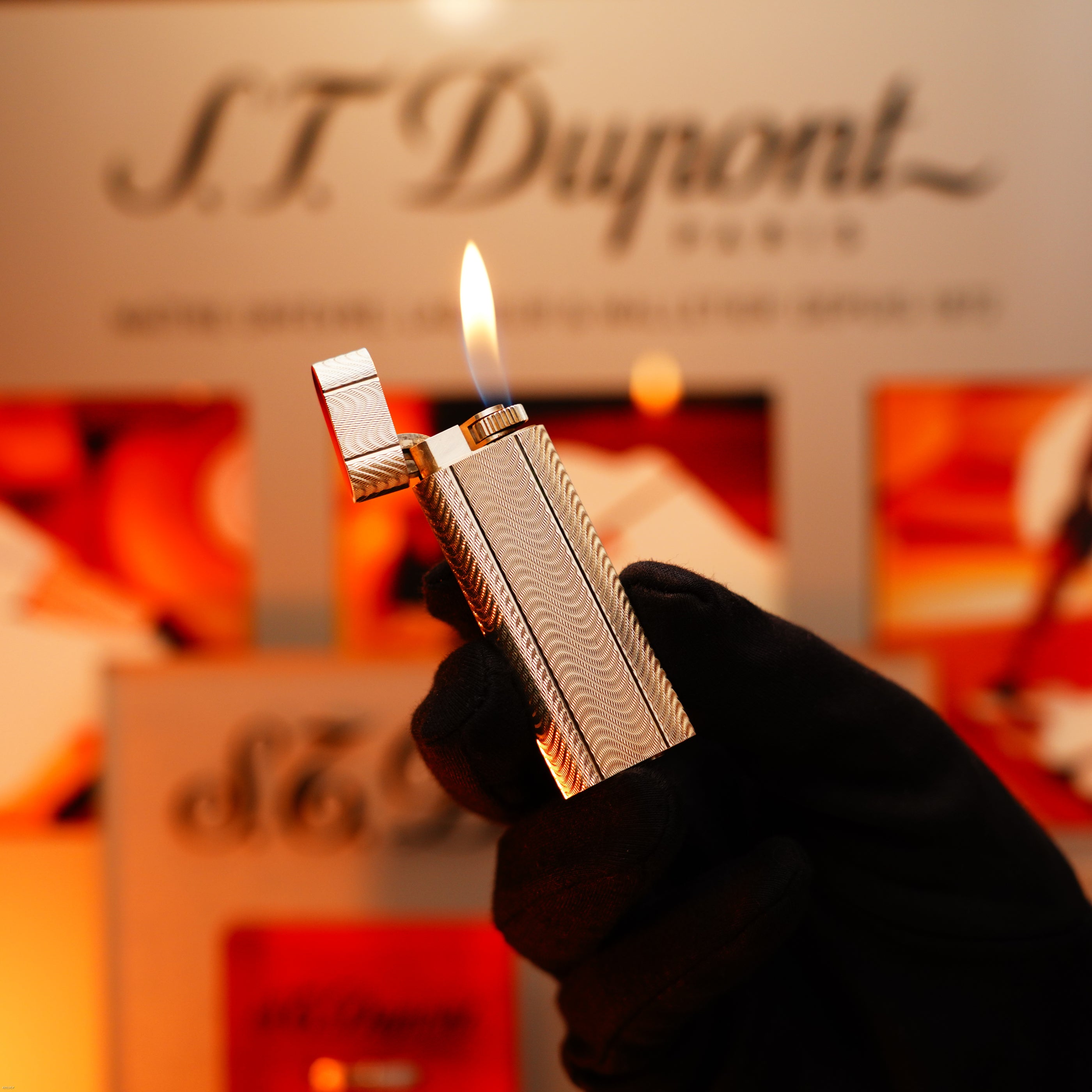 A gloved hand lights the Vintage 1970 Early Cartier Production Heavy Silver Rare Guilloché Pattern Lighter from Lucy Lighter, its orange flame glowing—timeless design and quality reflected in a branded display behind.