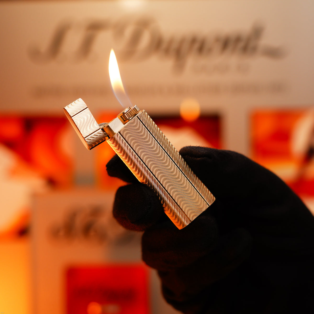 A gloved hand holds a Vintage 1970 Early Cartier Production Heavy Silver Rare Guilloché Pattern Lighter by Lucy Lighter, open with a visible flame, set against a blurred display to highlight its timeless elegance and quality.