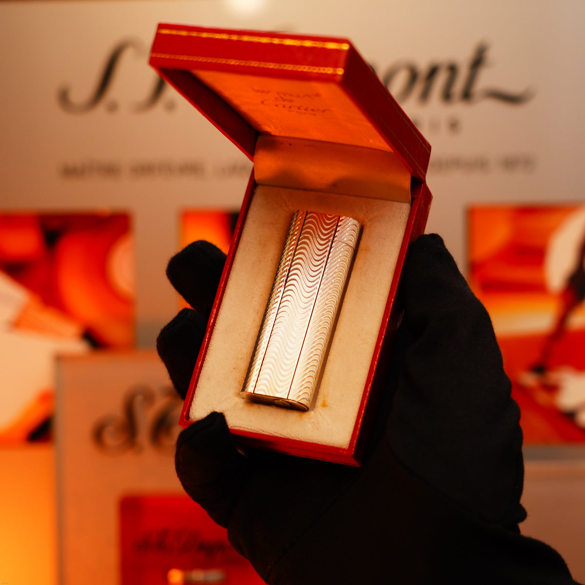 A gloved hand holds an open red box displaying a Vintage 1970 Early Cartier Production Heavy Silver Rare Guilloché Pattern Lighter by Lucy Lighter, set against abstract art and the Lucy Lighter logo—a mark of timeless design and quality.